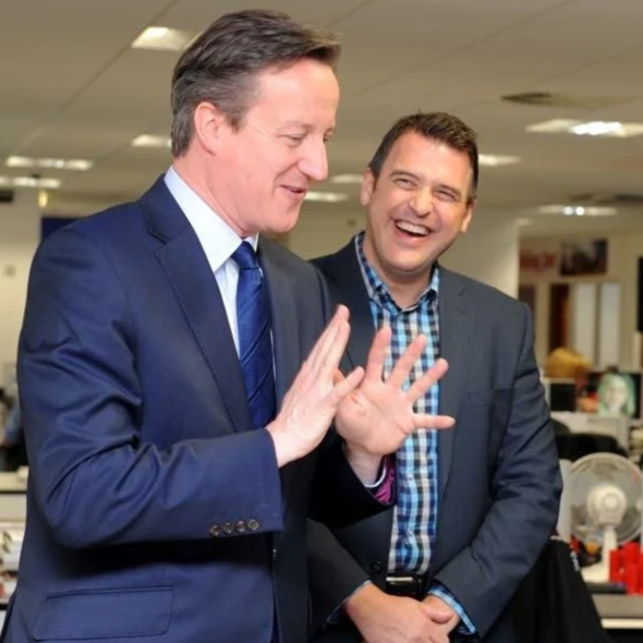 Two men in business suits laughing and smiling indoors, one with his hand raised as if gesturing or mid-conversation.