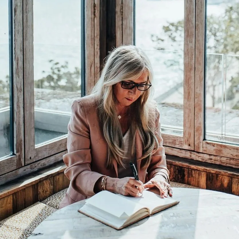 A woman with long blonde hair and glasses sitting at a round marble table, writing in a notebook, next to large windows with wooden frames, in a cozy, rustic setting.