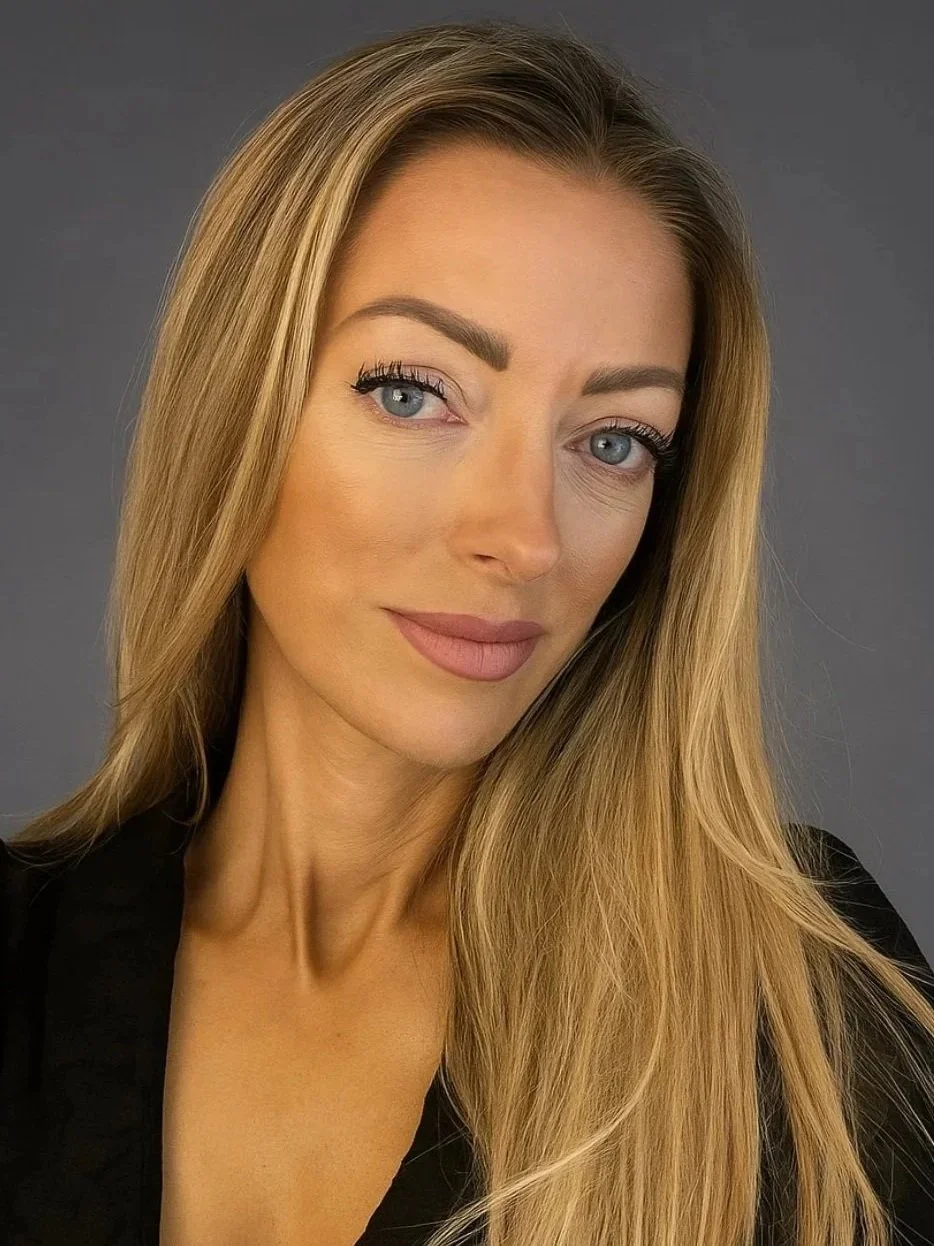 A woman with long blonde hair, blue eyes, and light makeup, wearing a black top, posing against a gray background.