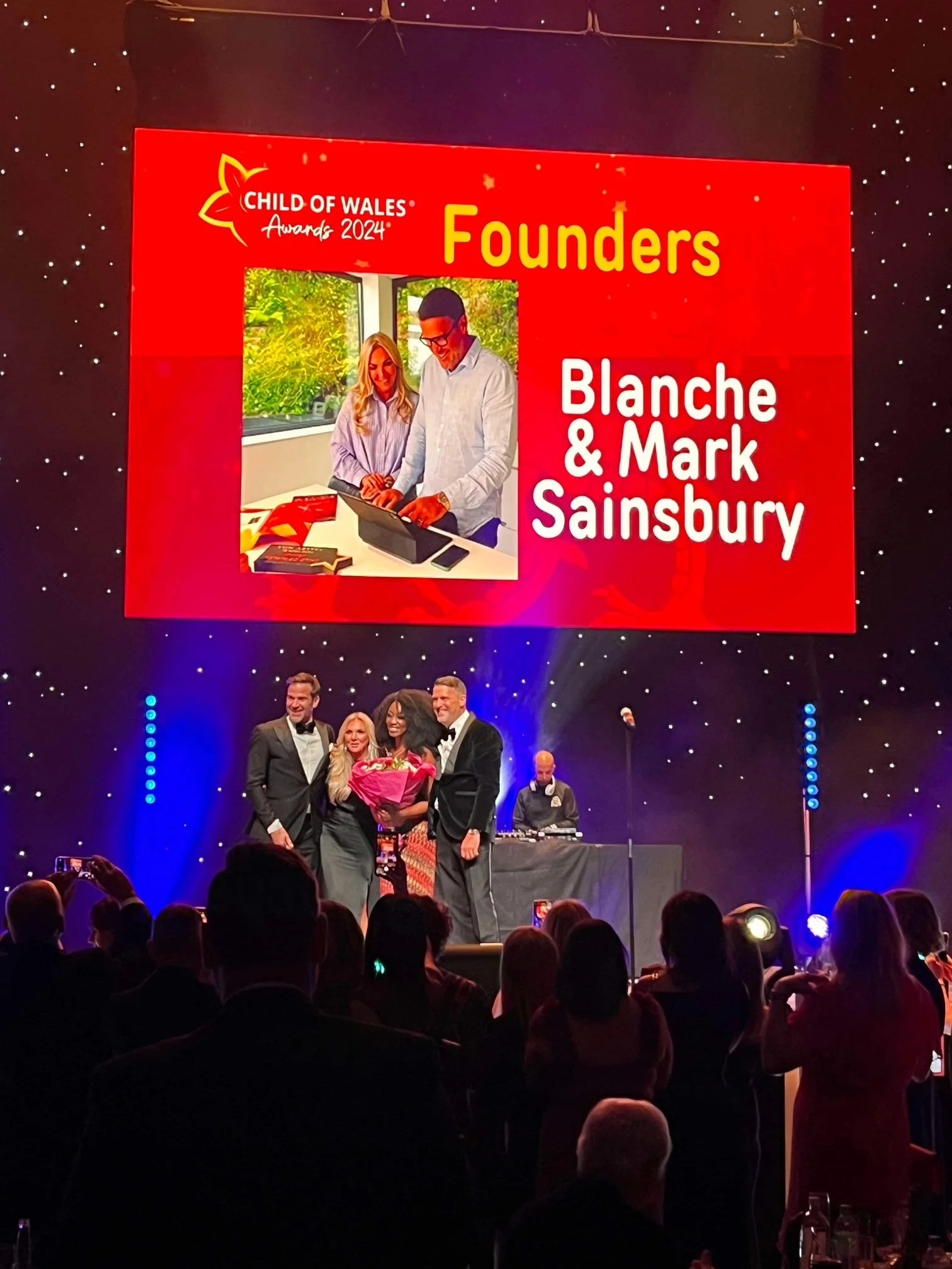 An award ceremony on stage with a large screen in the background. The screen displays the Child of Wales Awards 2024, highlighting founders Blanche and Mark Sainsbury. Four people are on stage, three dressed in tuxedos and one in a dress, with someone holding a bouquet of flowers.