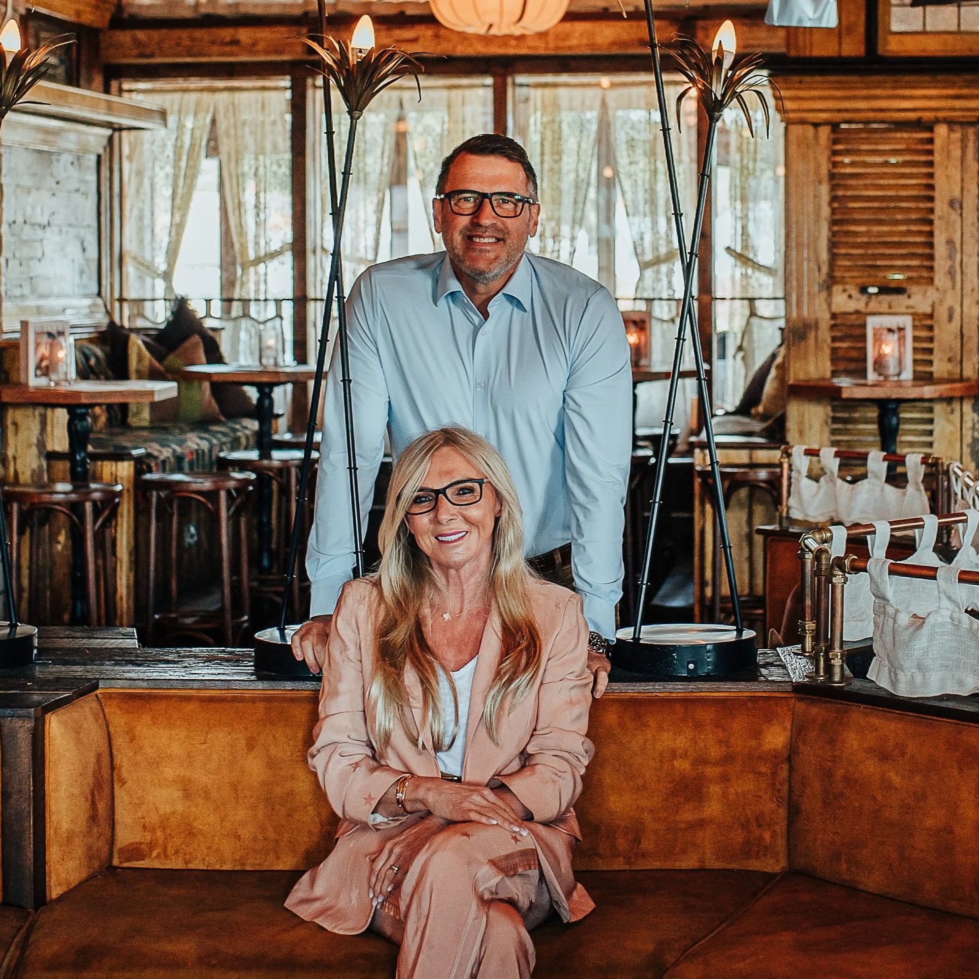 A man in a light blue shirt and glasses standing in a rustic wooden restaurant or cafe, smiling, with a woman in a pink jacket sitting at a table in front of him, and warm lighting and wooden decor in the background.