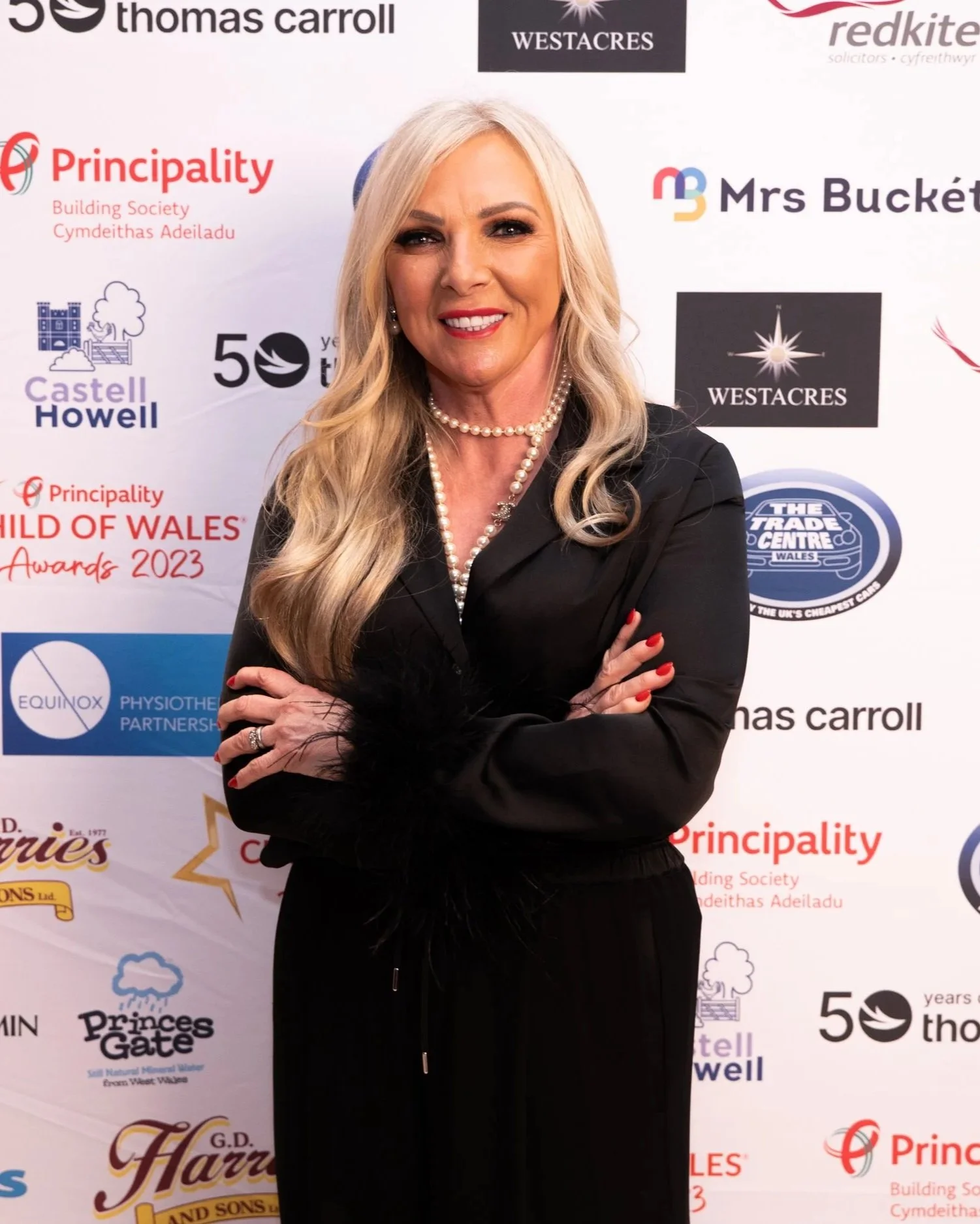 A woman with long blonde hair wearing a black outfit and pearl necklace, standing in front of a backdrop with various sponsor logos and text.