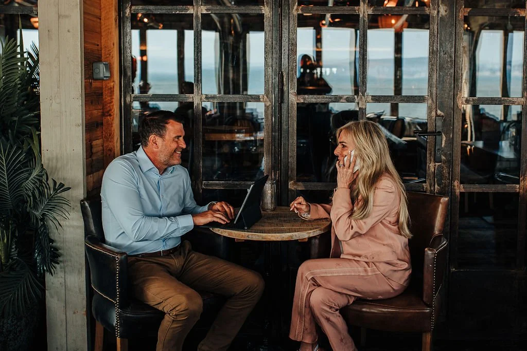Man and woman sitting at a small round table in a cozy restaurant, talking and smiling, with the man using a laptop and the woman talking on a cell phone, wooden walls, large glass window, warm ambient lighting.