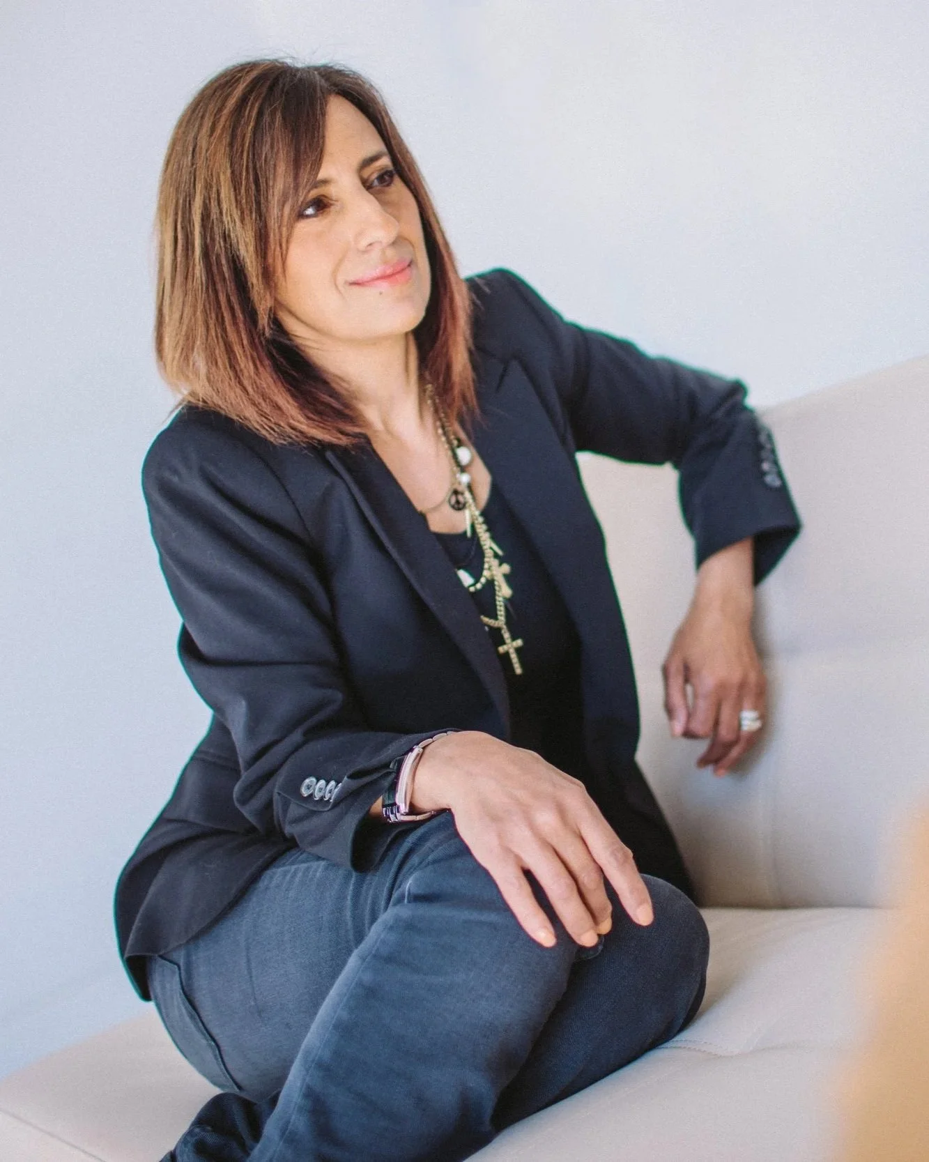 A woman with shoulder-length reddish-brown hair sitting on a beige sofa, wearing a black blazer and dark jeans, accessorized with a necklace with multiple crosses, a watch, and a bracelet, looking thoughtfully to her left.