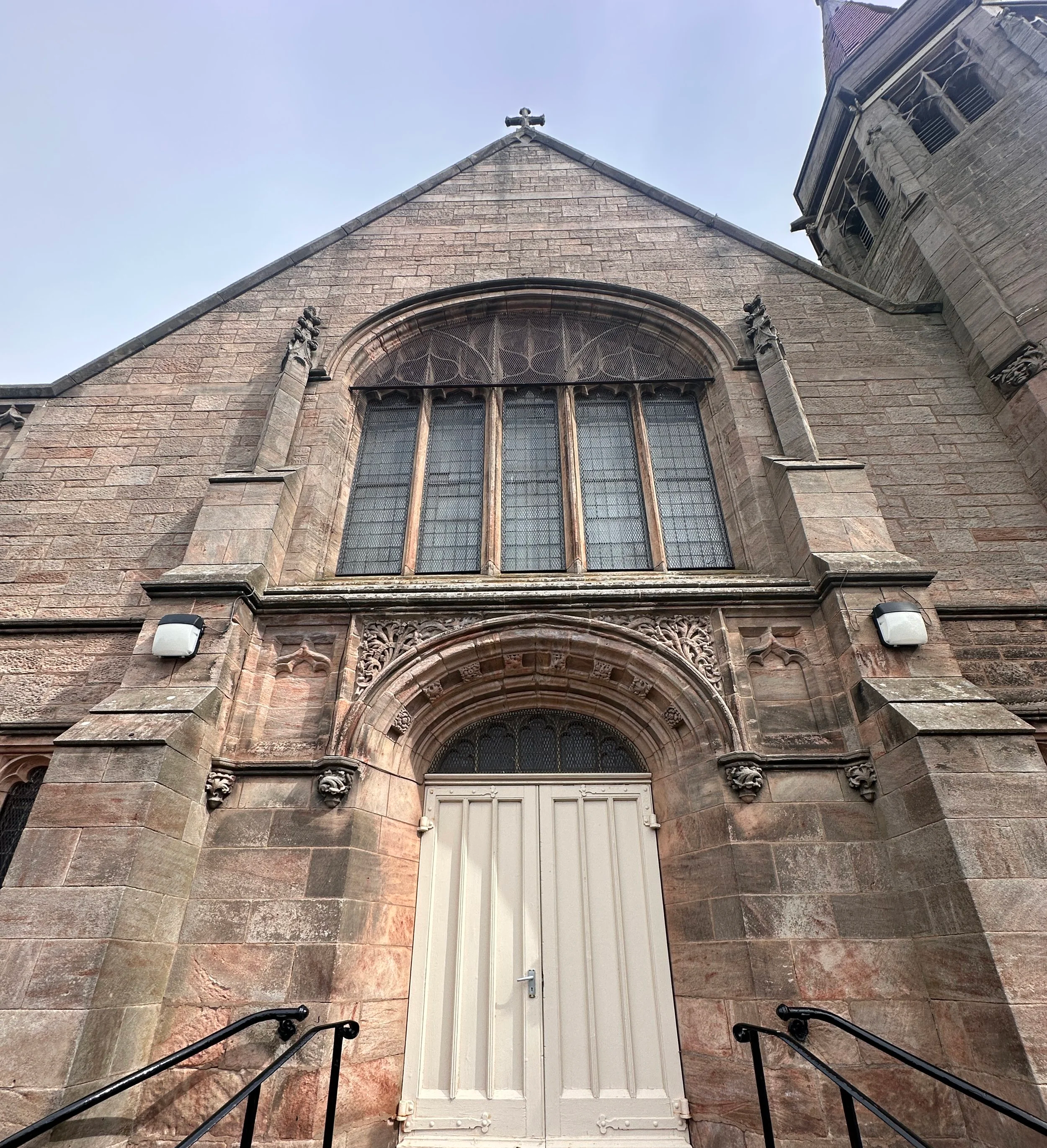Front view of a historic stone church with gothic architectural details, including decorative carvings and large stained glass windows, accessed by stone steps with black metal railings.