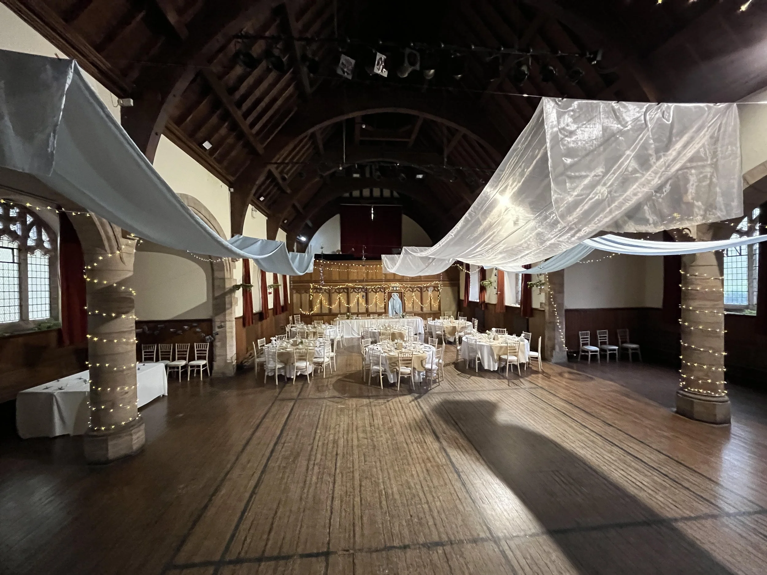 A decorated banquet hall with white drapes and string lights, round tables with white tablecloths, and wooden chairs, inside a historic building with high arched wooden ceilings and windows.