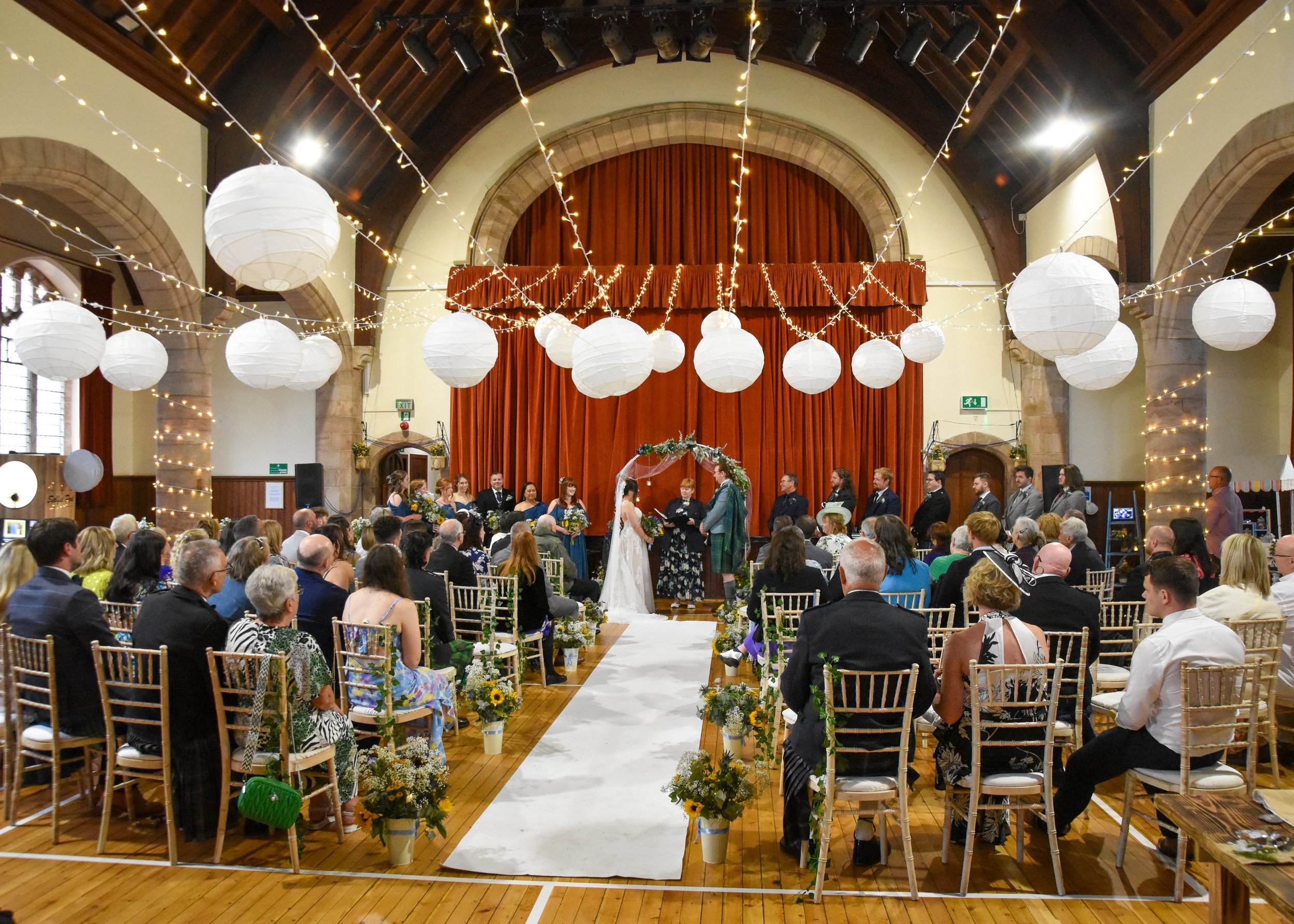 A wedding ceremony inside a decorated hall with hanging white lanterns and string lights, an arch with flowers at the altar, and guests seated in chairs.