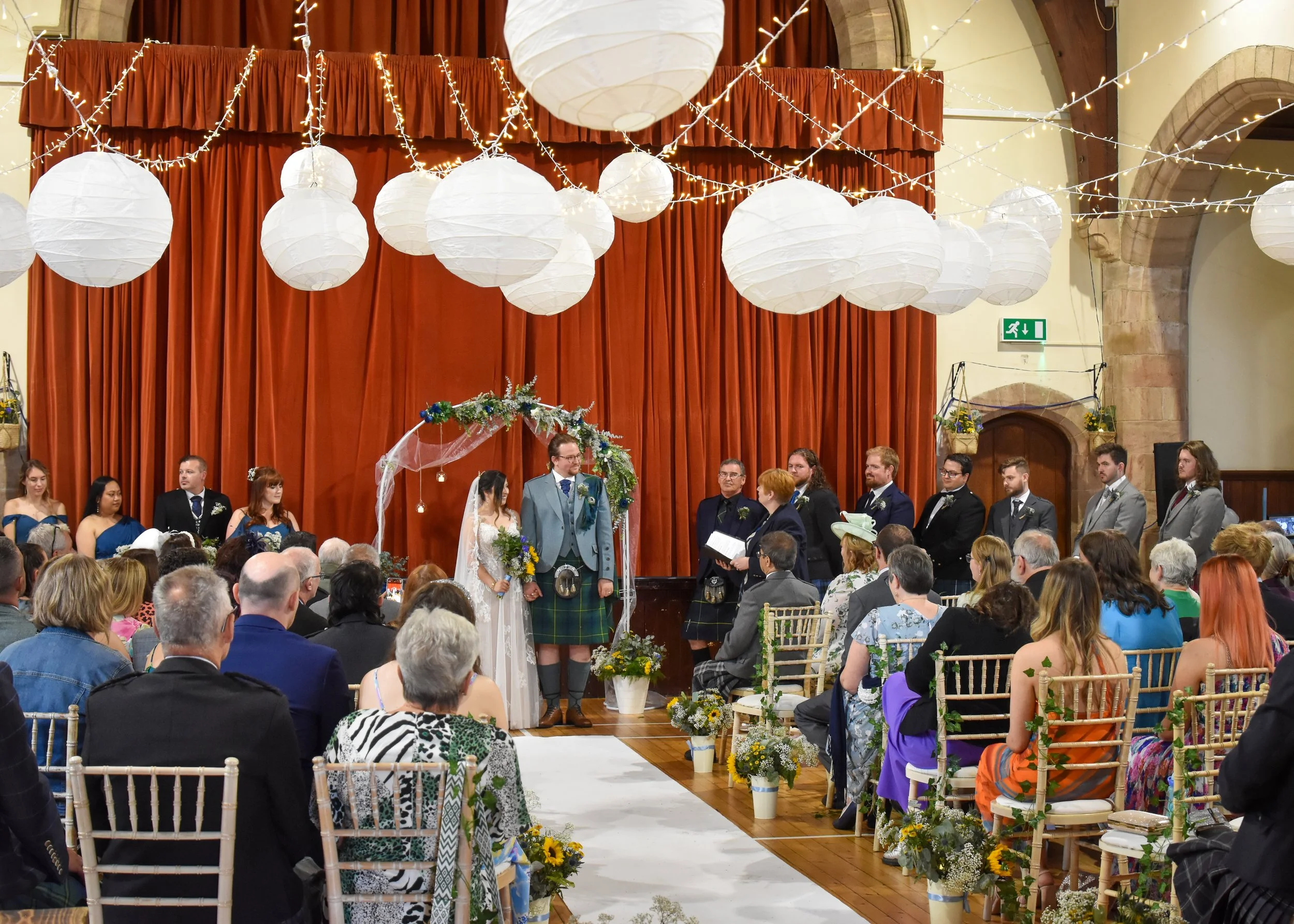 A wedding ceremony inside a decorated Crail Community Hall with white paper lanterns and string lights hanging from the ceiling, red curtains on the stage, and guests seated facing the couple.