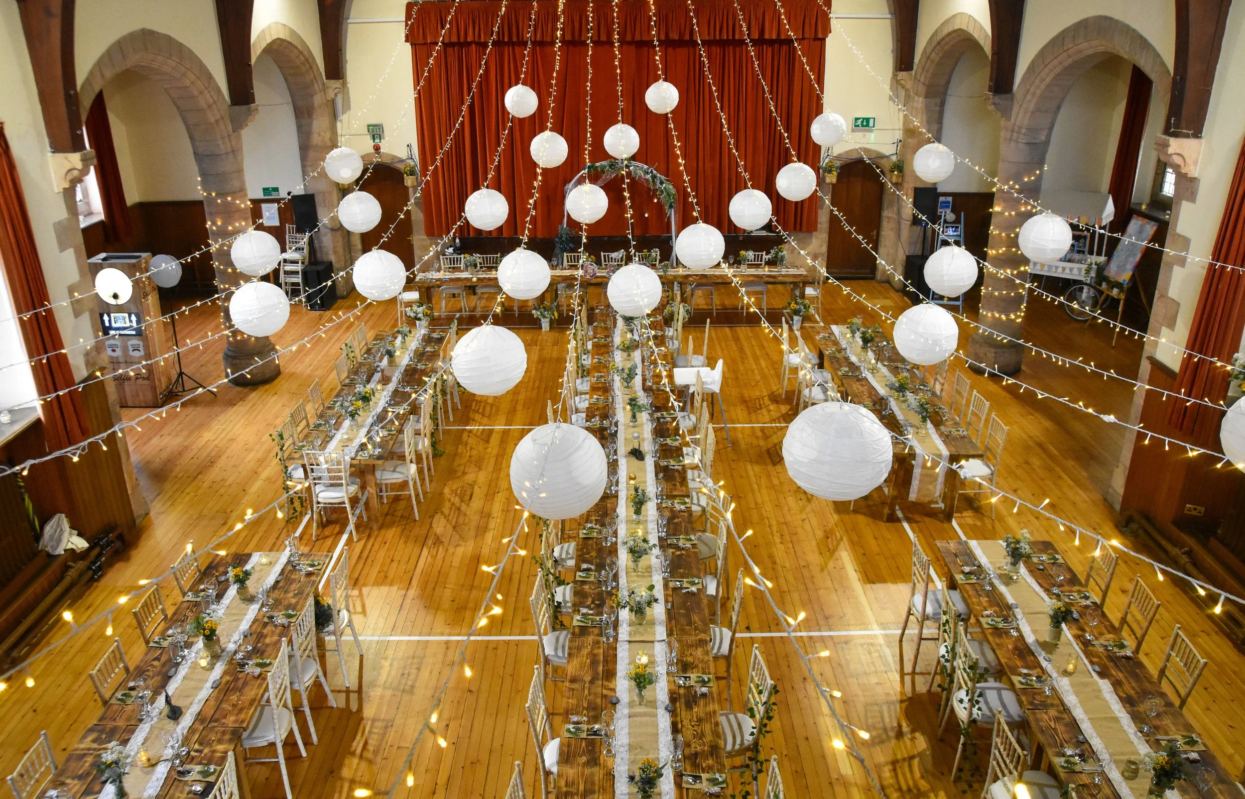 Wedding reception setup in a large decorated hall with long tables, white chairs, floral centerpieces, hanging white paper lanterns, and string lights.