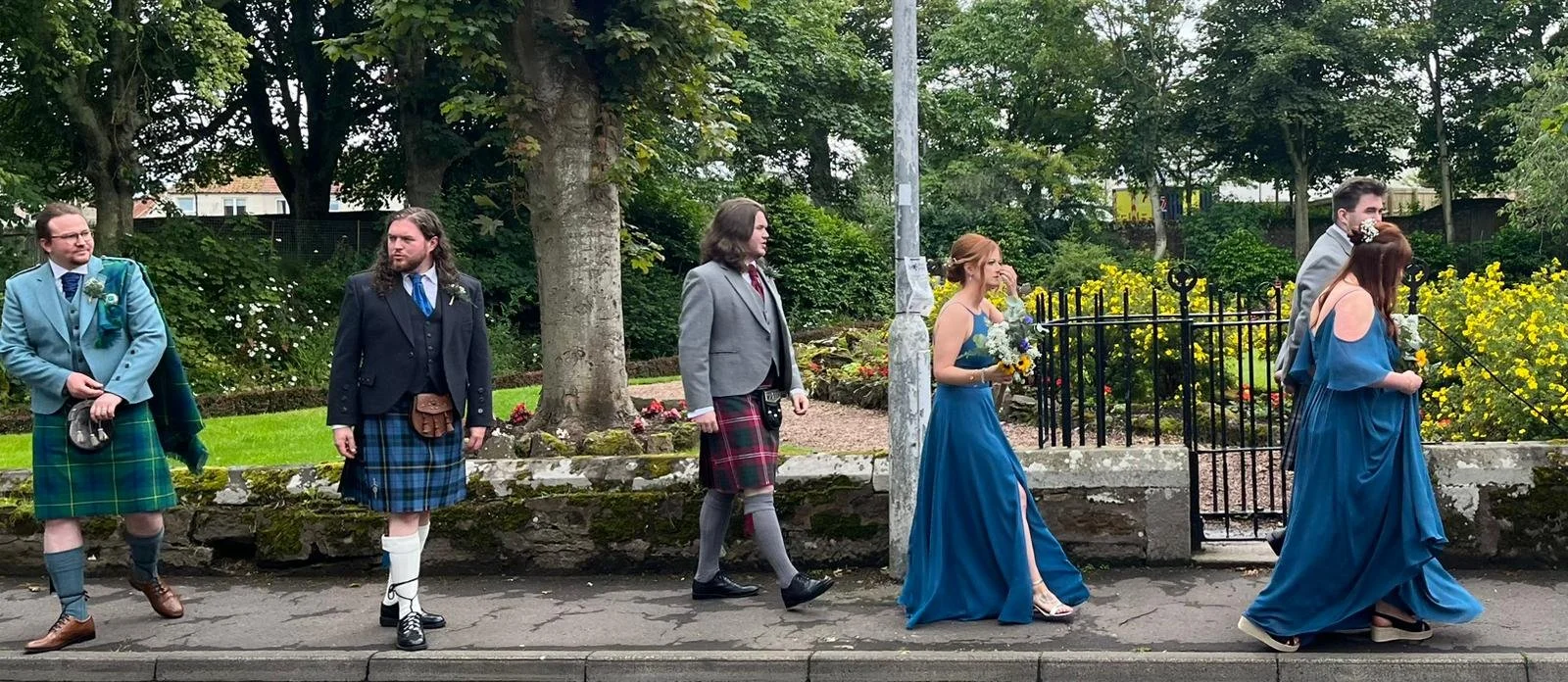 Group of people dressed in formal and traditional Scottish attire walking outdoors on a sidewalk with greenery and flowers behind them.