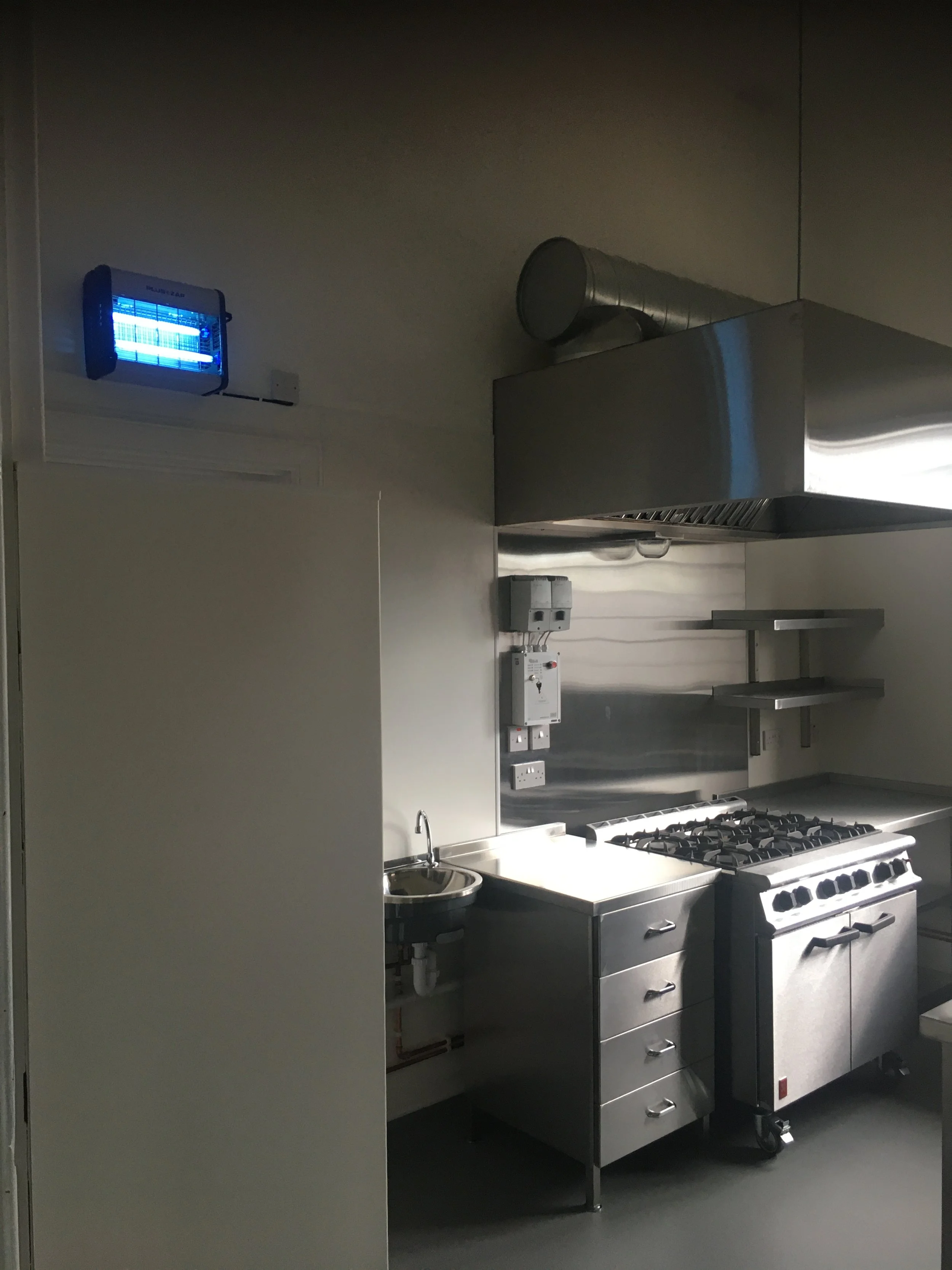 Commercial kitchen with stainless steel appliances including a stove, countertop, and ventilation hood. There is a small sink, electrical outlets, and a blue insect trap on the wall.