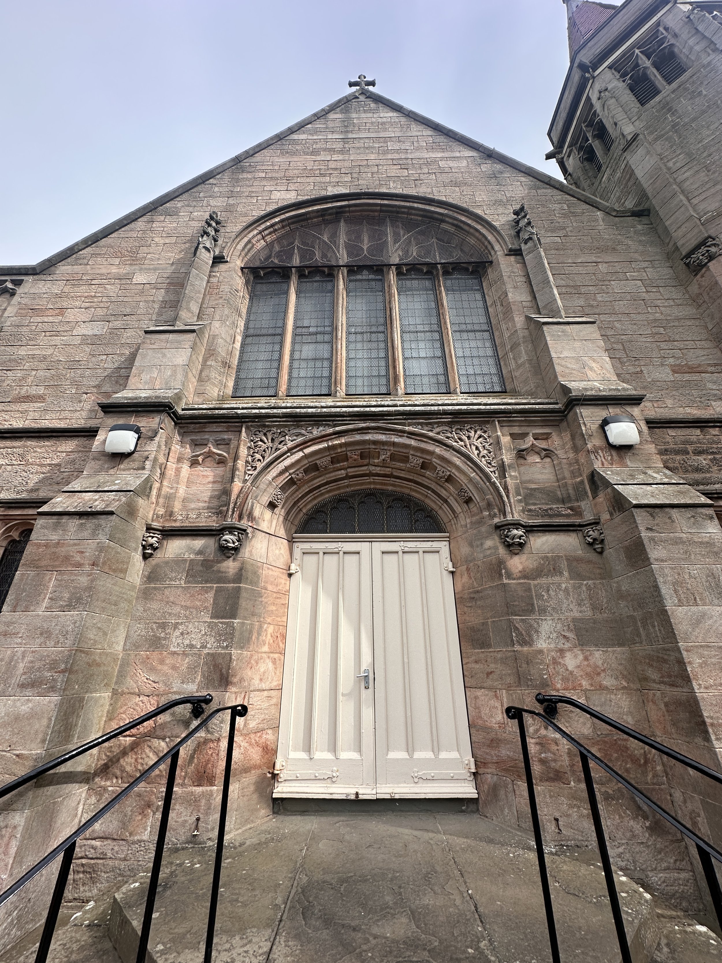 Front view of a stone church entrance with a white wooden door, arched stained glass window above, and decorative stonework on the facade. Black metal railings lead up to the door, and a cross is visible at the top of the building.