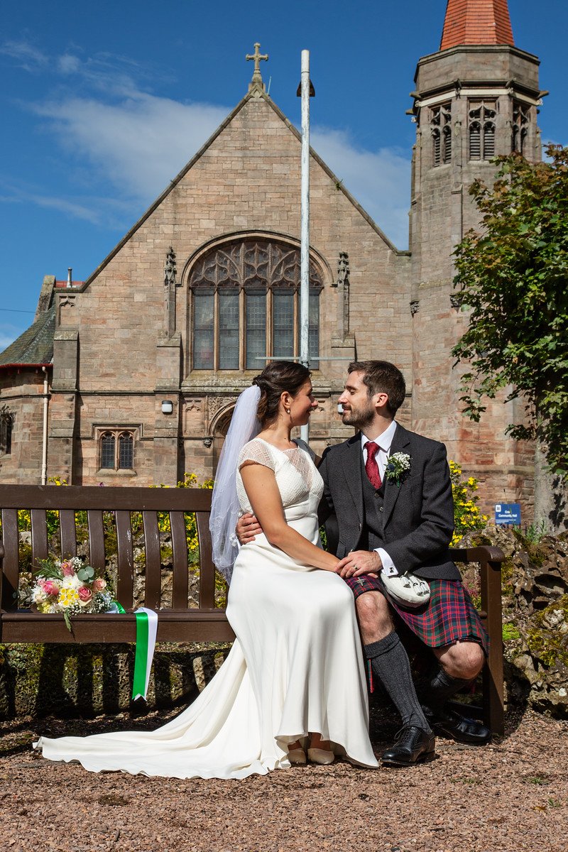 A bride and groom sitting closely on a wooden bench in front of a church, with a bouquet of flowers on the side of the bench. The bride is wearing a white wedding dress with a veil, and the groom is wearing a dark suit with a red tie and a kilt. They