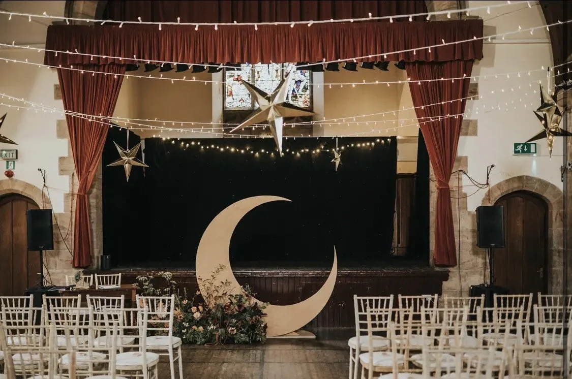 Interior of a decorated Crail Community Hall stage with a stage featuring a large crescent moon and floral arrangement, red curtains, string lights, and hanging star and moon decorations.