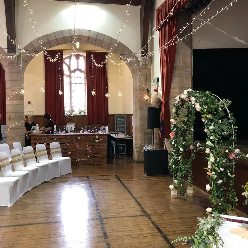 Interior of a decorated wedding or event hall with chairs, floral arch, string lights, and a serving table.