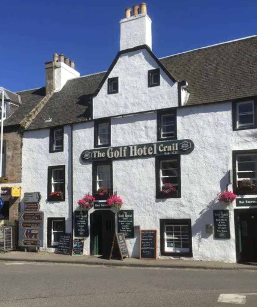 A white building with black trim named "The Golf Hotel Crail," established in 1821. It has several windows with flower boxes filled with pink flowers, a sign above the door, and multiple chalkboards and signs near the entrance. The sky is clear and blue.