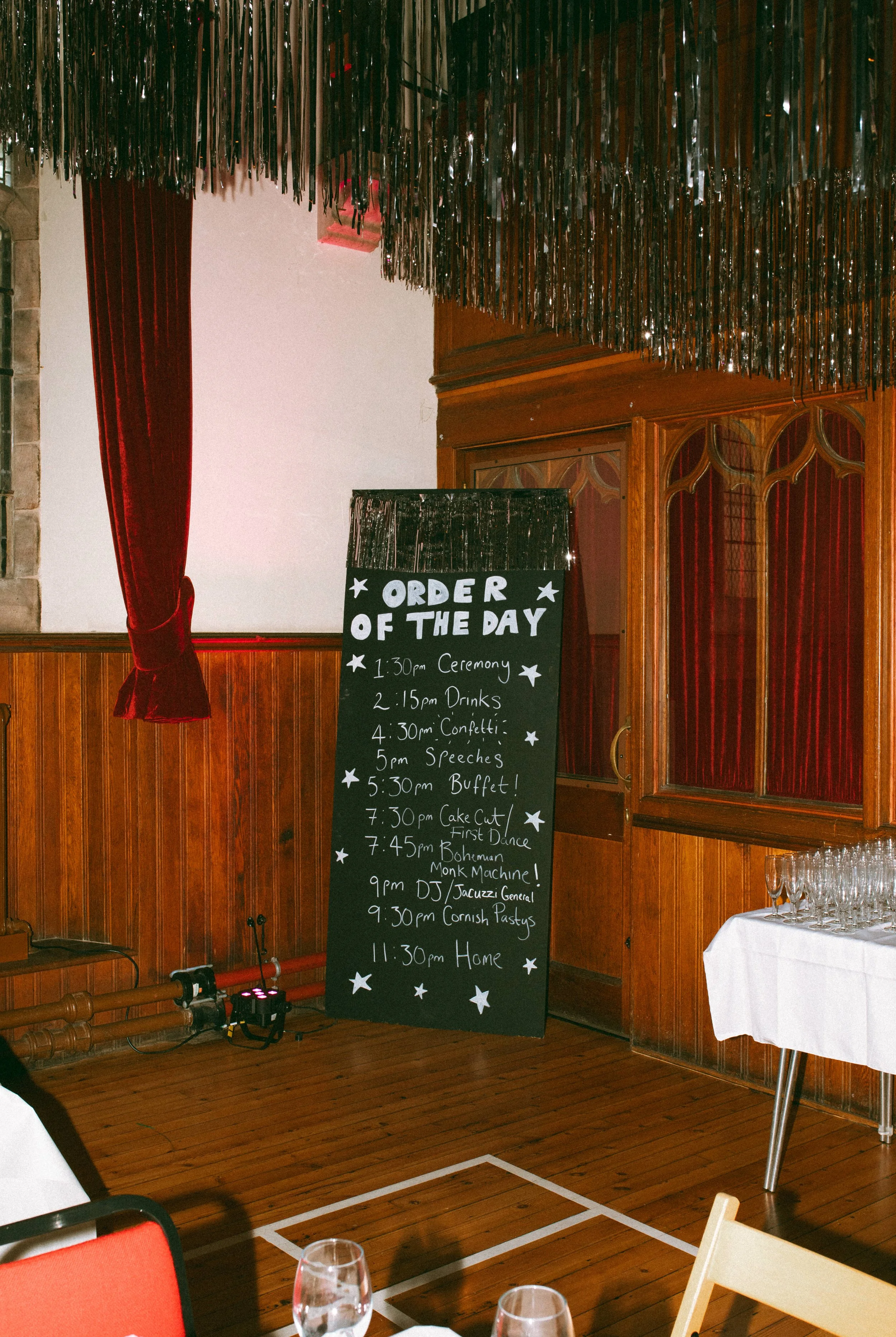 A black chalkboard with white handwritten text listing the schedule of events for a celebration or party, including times for ceremony, drinks, confetti, speeches, buffet, cake cut, dancing, karaoke, and home. The board is placed in a wooden-paneled 