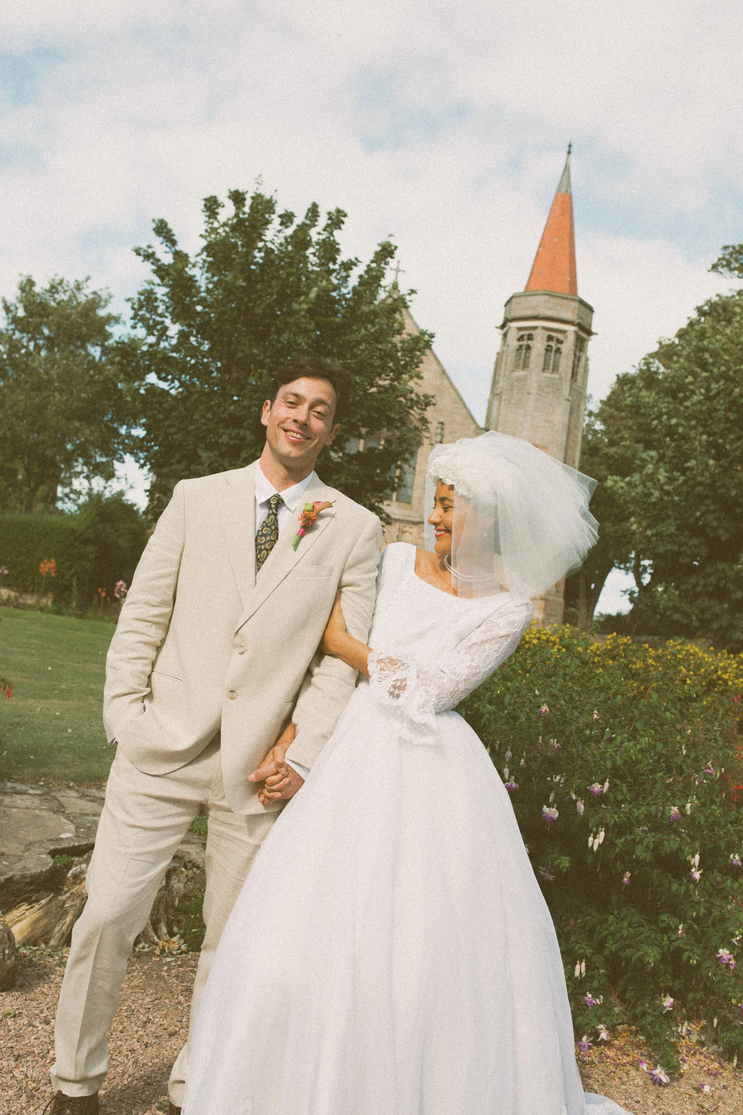 A happy couple outdoors in wedding attire holding hands, with a church and trees in the background.