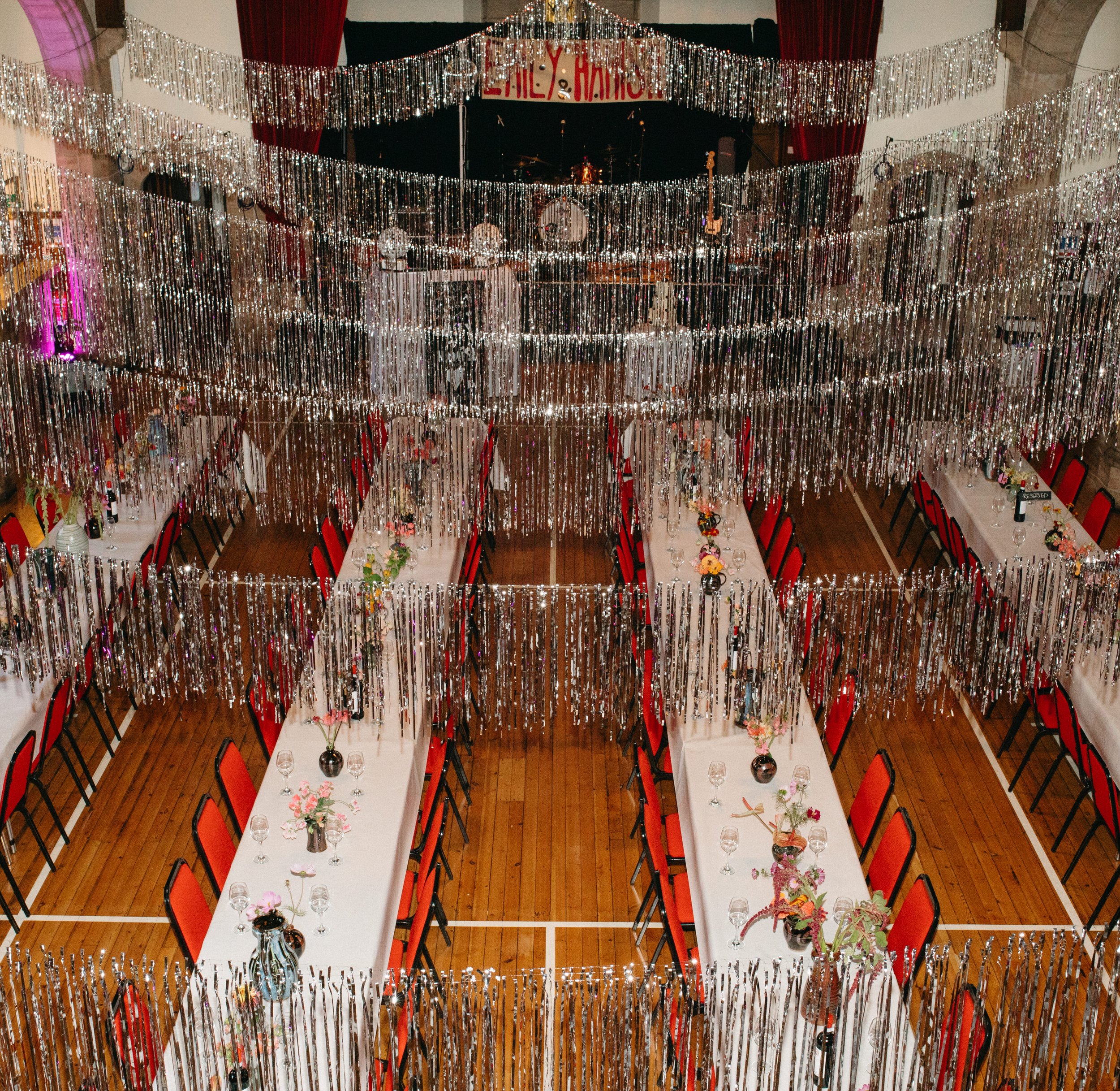 A banquet hall decorated with silver streamers hanging from the ceiling and a stage at the back with a drum set and microphones, with tables set for a celebration with floral centerpieces and place settings.