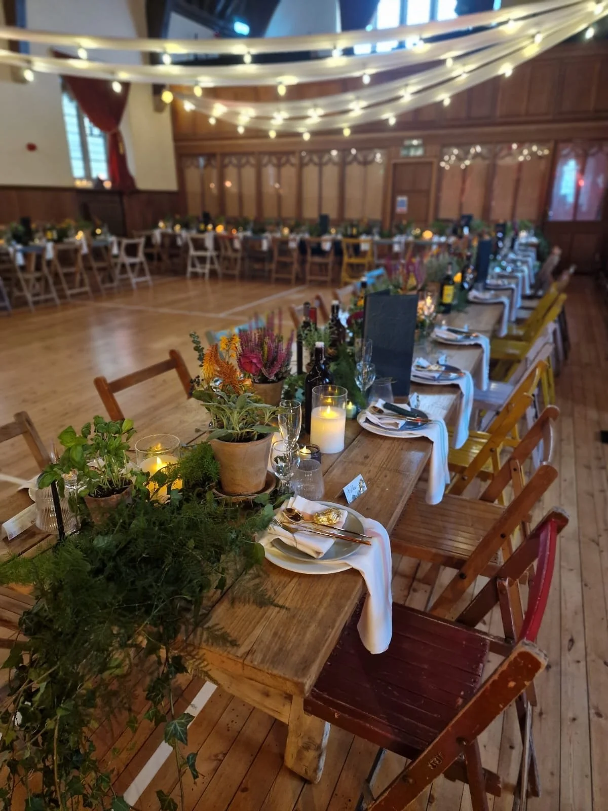 Indoor event space decorated with string lights and draped fabric, featuring long wooden tables set with flowers, candles, and dining ware for a celebration or gathering.