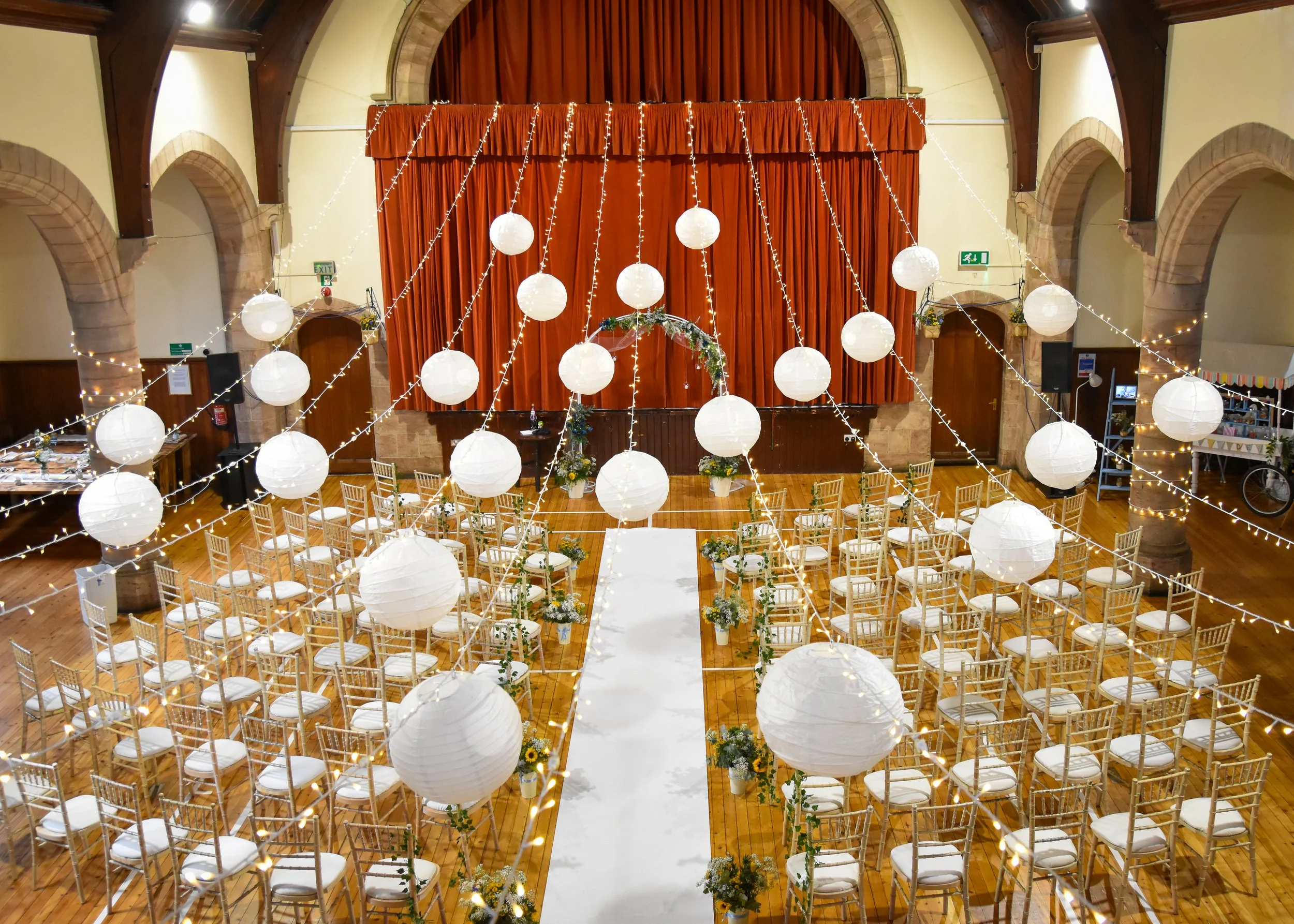 A decorated wedding ceremony hall with white paper lanterns and string lights hanging above rows of white chairs with gold frames, a red curtain backdrop, and floral arrangements.