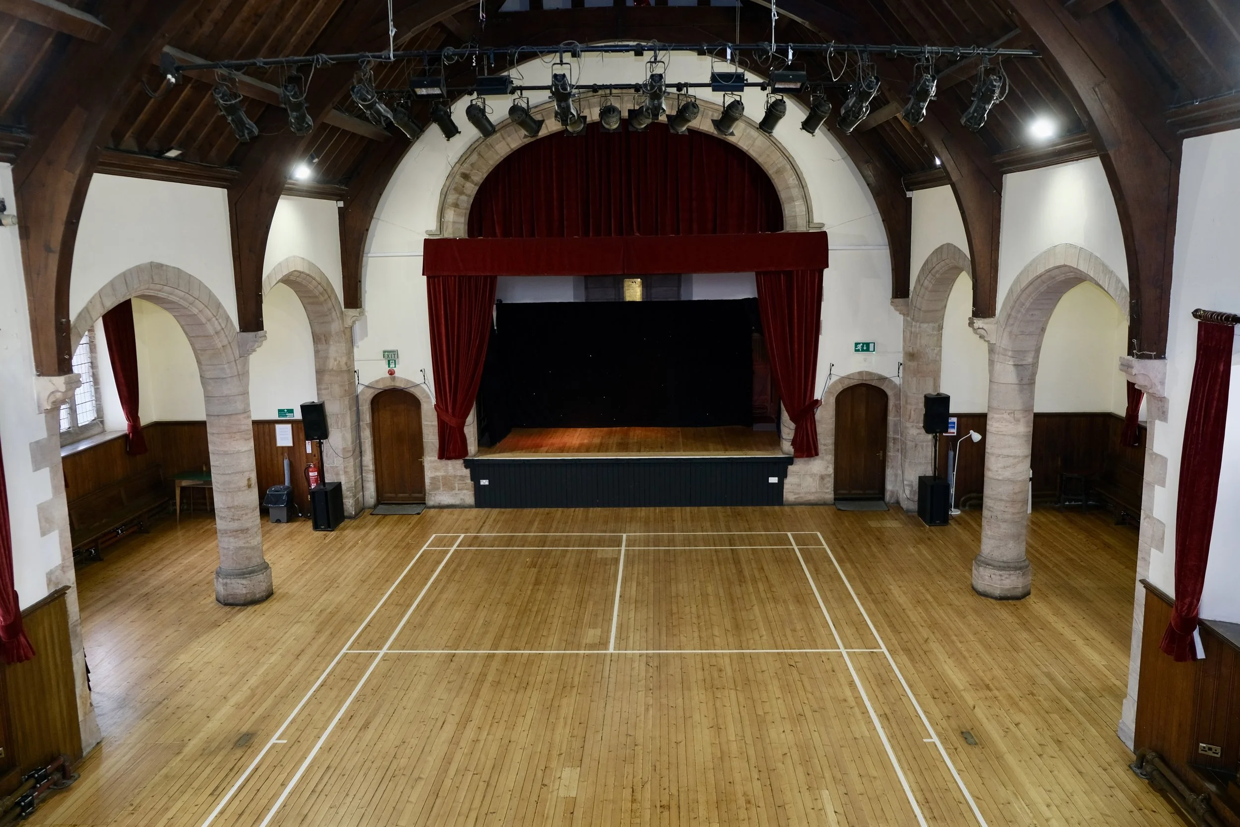 Empty gymnasium with a basketball court, wooden floors, high arched ceiling, stone columns, red stage curtains, and stage lights.