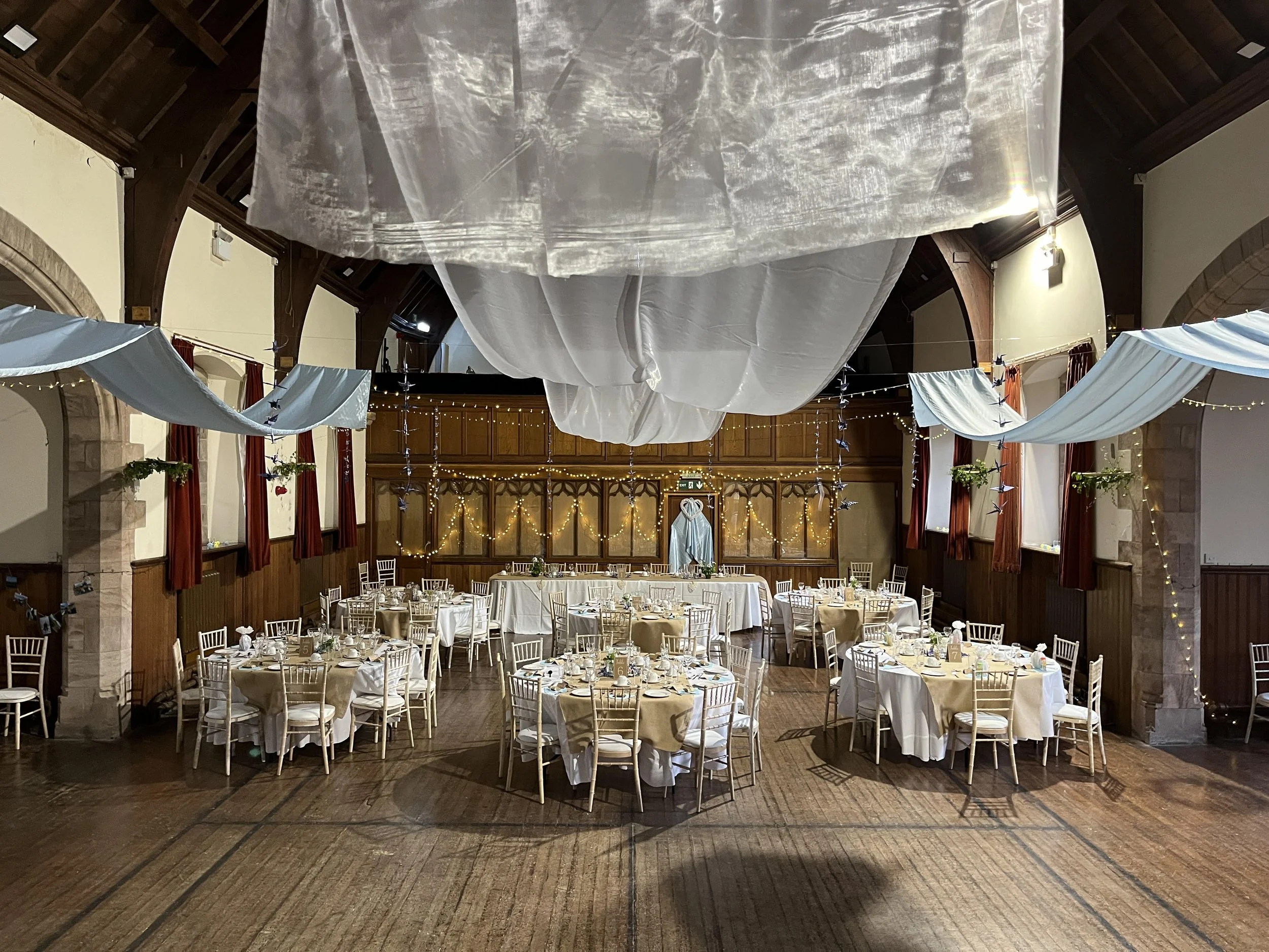 Elegant banquet hall decorated for a wedding reception with round tables covered in white and beige tablecloths, surrounded by white chairs, with hanging sheer fabric and string lights overhead.