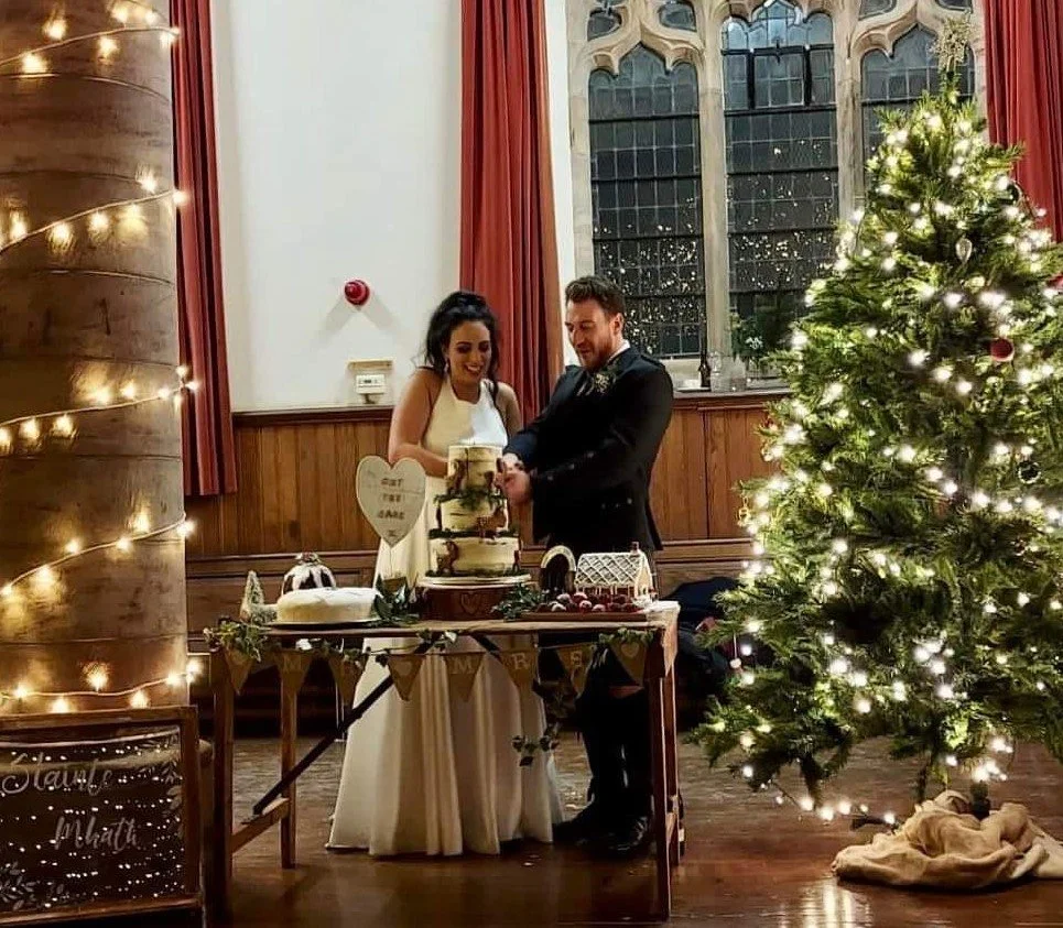 A bride and groom cutting a wedding cake at a Christmas-themed reception inside a church with a decorated Christmas tree and warm lighting.
