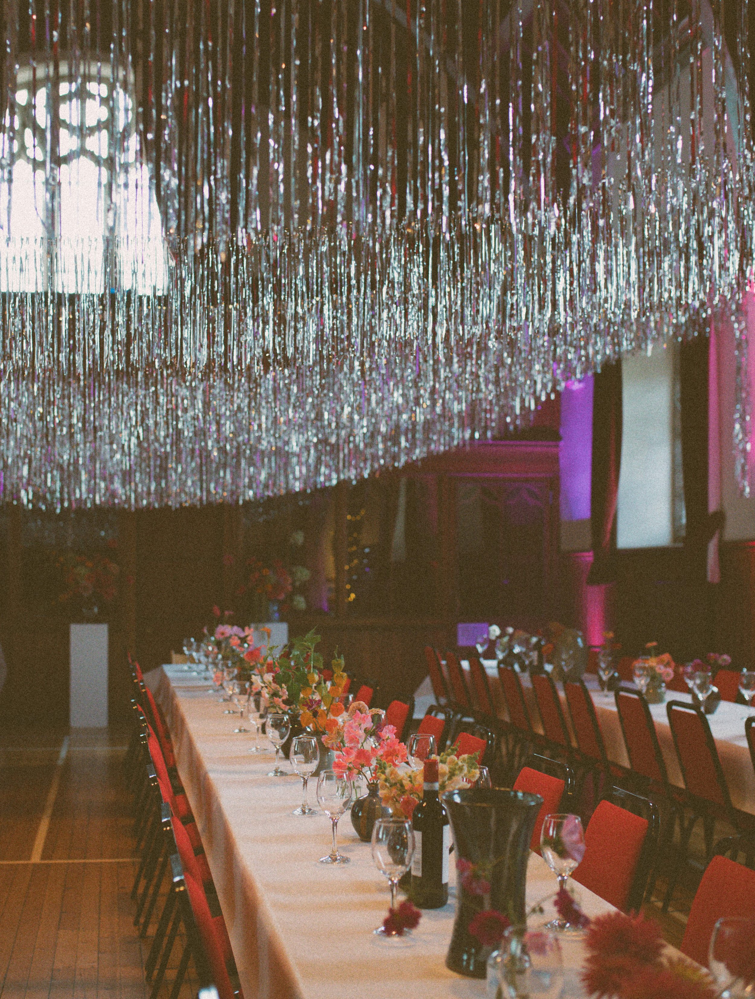 Decorative event banquet hall with long dining table set with floral centerpieces, glassware, and bottles, under a shimmering chandelier with metallic streamers, and pink lighting accents.