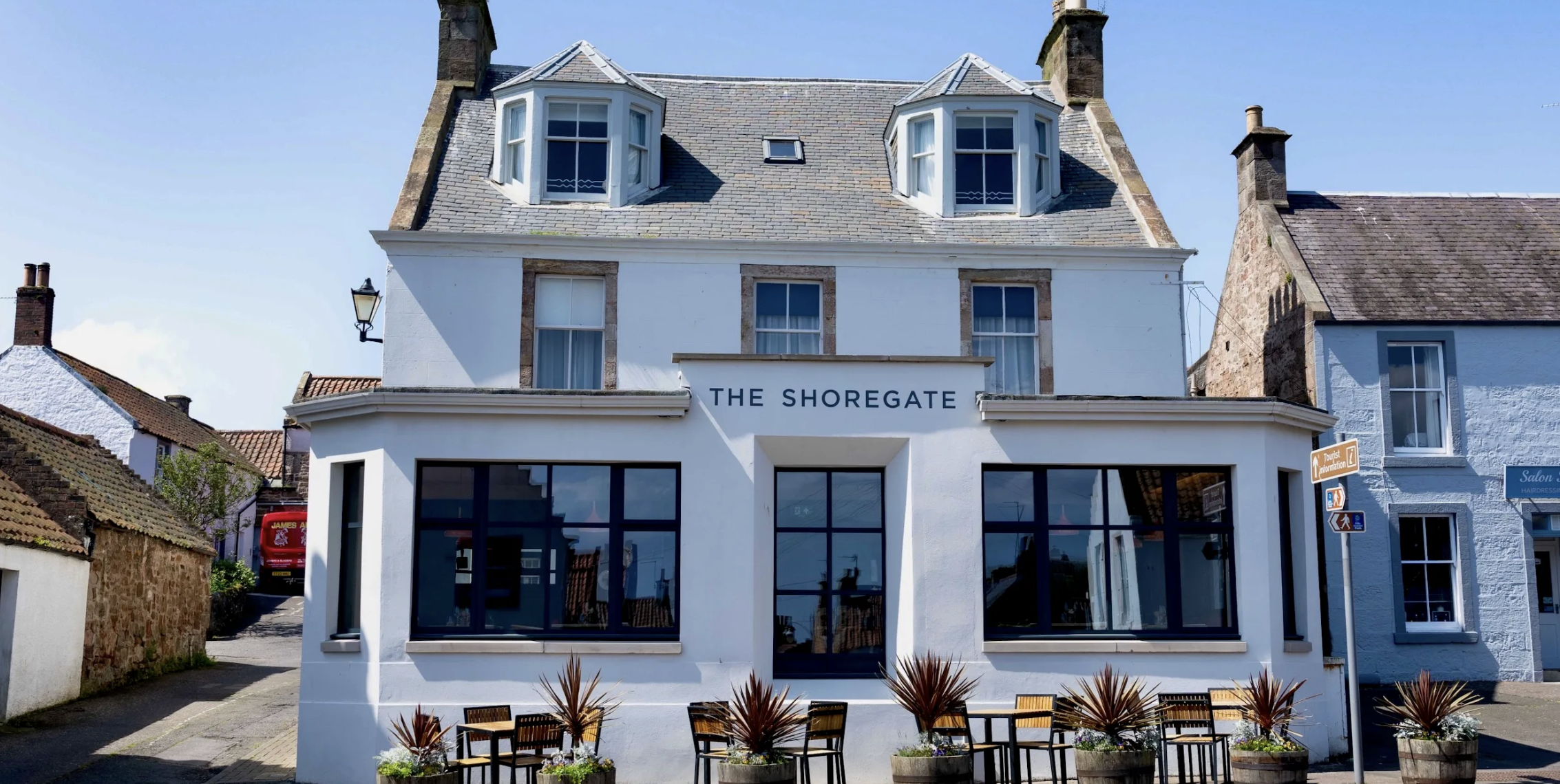 A white building named 'The Shoregate' with large glass windows on the ground floor and two dormer windows on the roof, located in a small town with other stone and white houses and a sign indicating a hairdresser nearby.