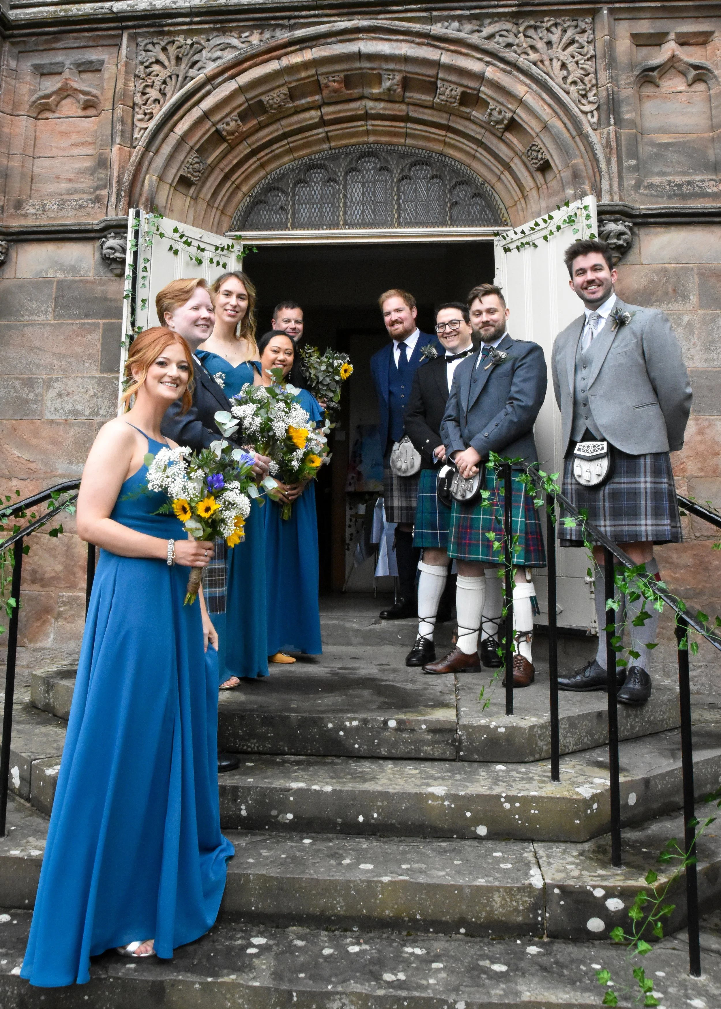 A group of people dressed in formal attire standing on the steps of a church. The women are in blue dresses holding bouquets, and the men are in suits with kilts. Some are smiling for the camera.