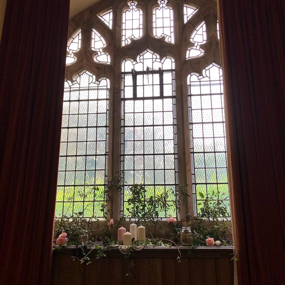 Inside a historic building with a large ornate stained glass window, decorated with candles, flowers, and greenery on the wooden window sill.