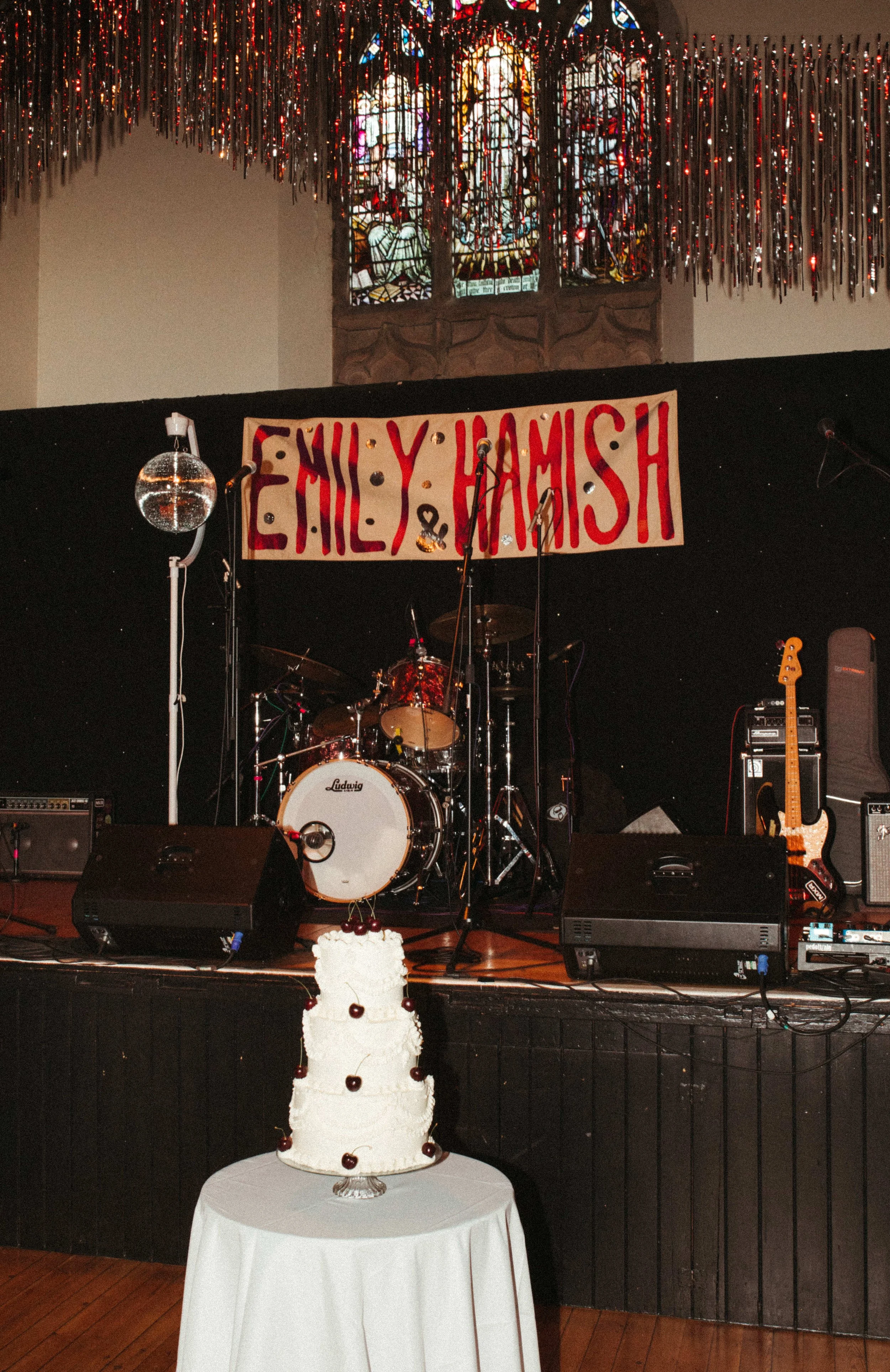 A concert stage decorated with a sign reading 'Emily & Hamish', a cake with cherries on a table in front, and musical instruments including a drum set and an electric guitar. A stained glass window and shiny hanging decorations are visible in the bac