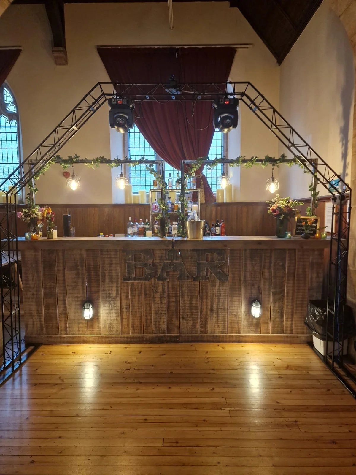 A rustic bar setup with a wooden counter, illuminated by hanging vintage-style light bulbs, decorated with flowers and greenery, with a metal frame arch overhead and candles in the background.