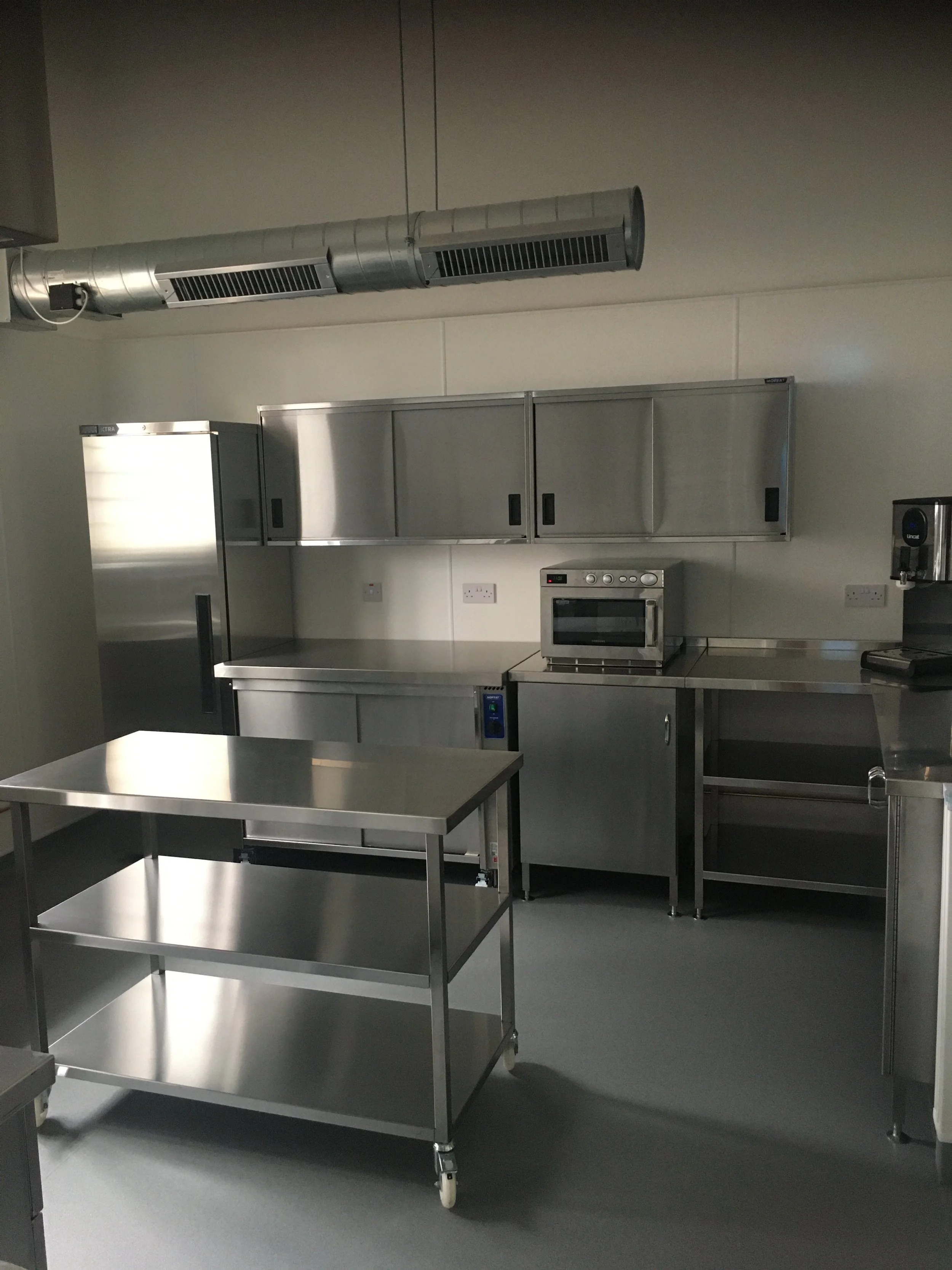 Empty commercial kitchen with stainless steel tables, cabinets, and appliances, including an oven and a refrigerator, in a room with gray walls and flooring.
