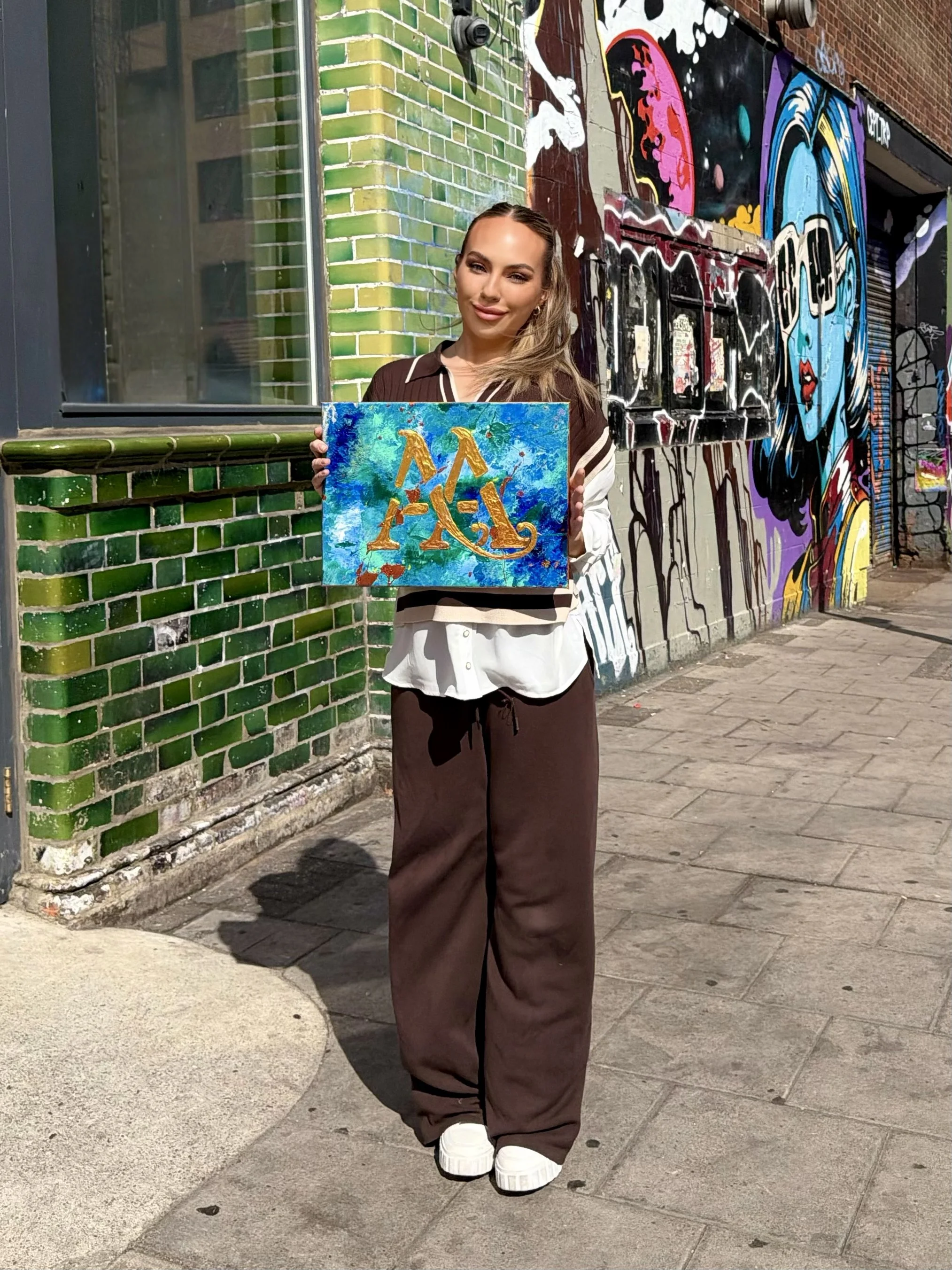 A woman with long blonde hair, wearing a brown and white shirt, brown pants, and white chunky shoes, standing on a sidewalk in front of a brightly colored graffiti wall, holding a painting with bold red, orange, and yellow letterings on a blue-green 