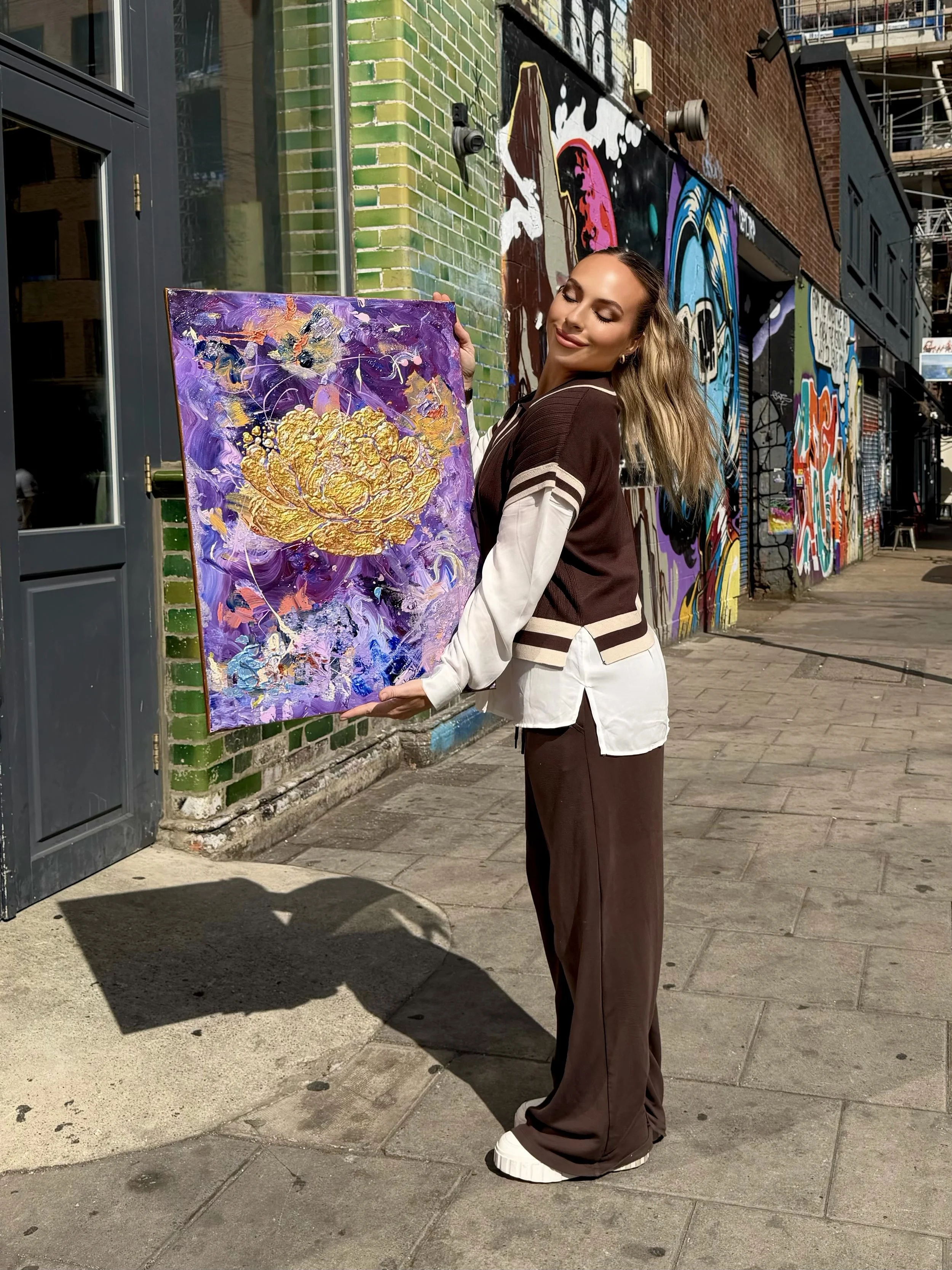 A woman with long blonde hair, dressed in a brown and white sporty jacket and brown wide-leg pants, stands outside on a city sidewalk, holding and looking at an abstract purple and gold painting. Behind her is a colorful mural painted on the brick wa