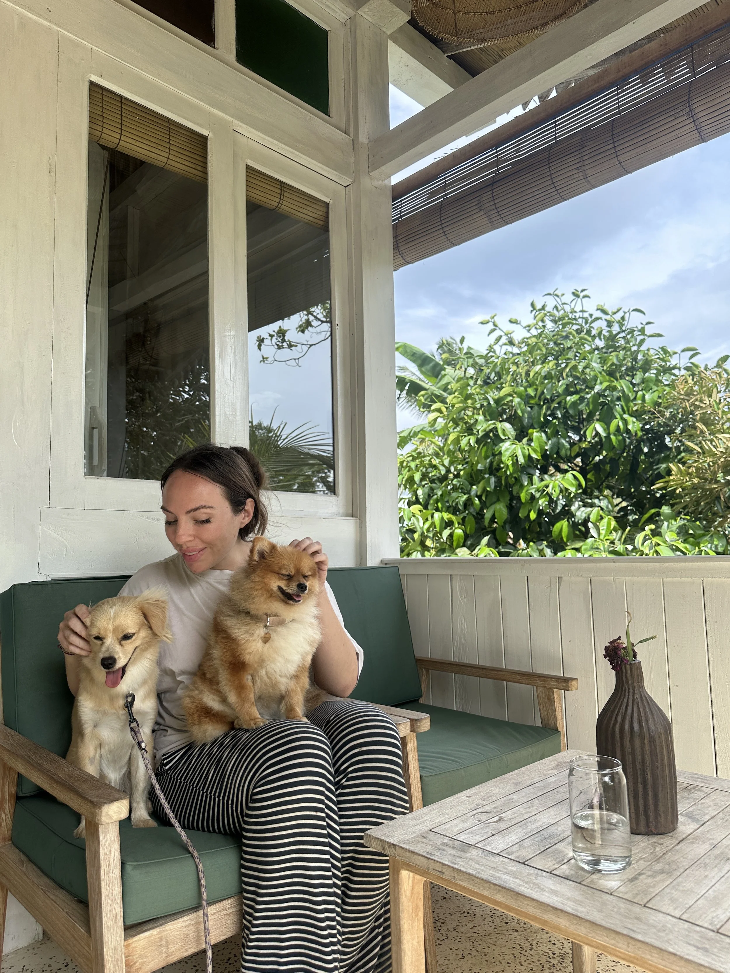 A woman sitting on a bench with two fluffy dogs, smiling and petting them, on a patio with a table with a glass of water and a decorative vase, surrounded by greenery and a bright sky.