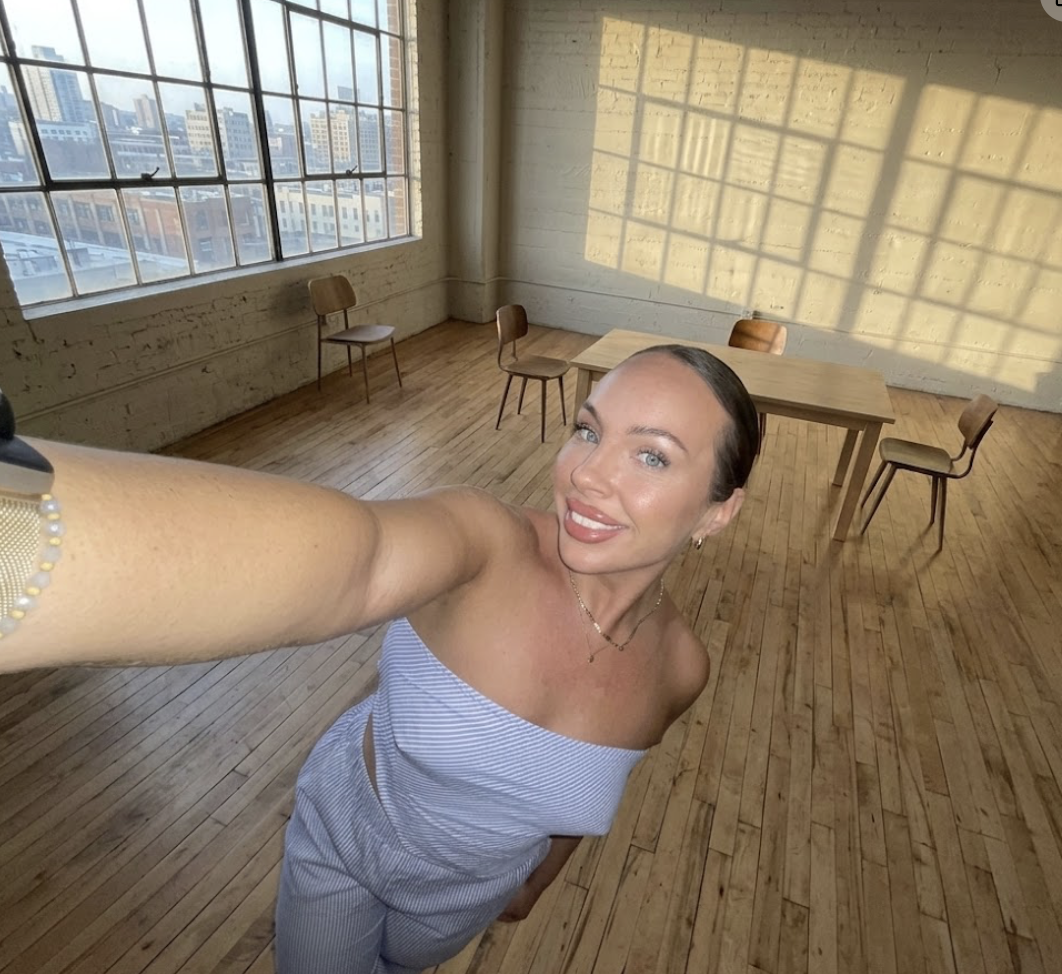 A smiling woman with dark hair and blue eyes taking a selfie in a spacious, bright room with large windows and wooden floors. The room is mostly empty except for a few wooden chairs and a table, with city skyline visible through the windows.