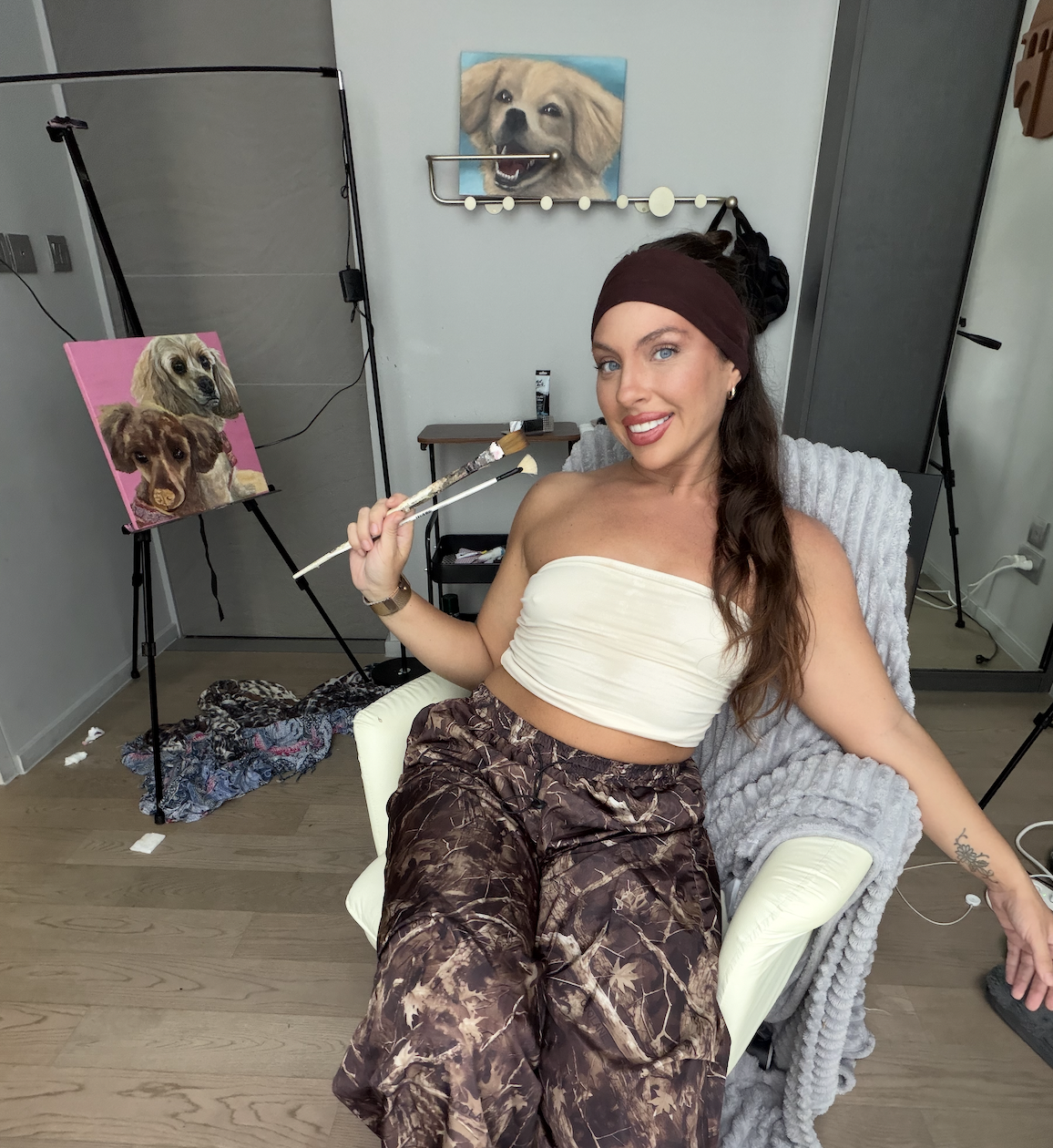 A woman sitting in a chair, holding paintbrushes, in a studio with dog portraits on easels and paintings of dogs on the walls.
