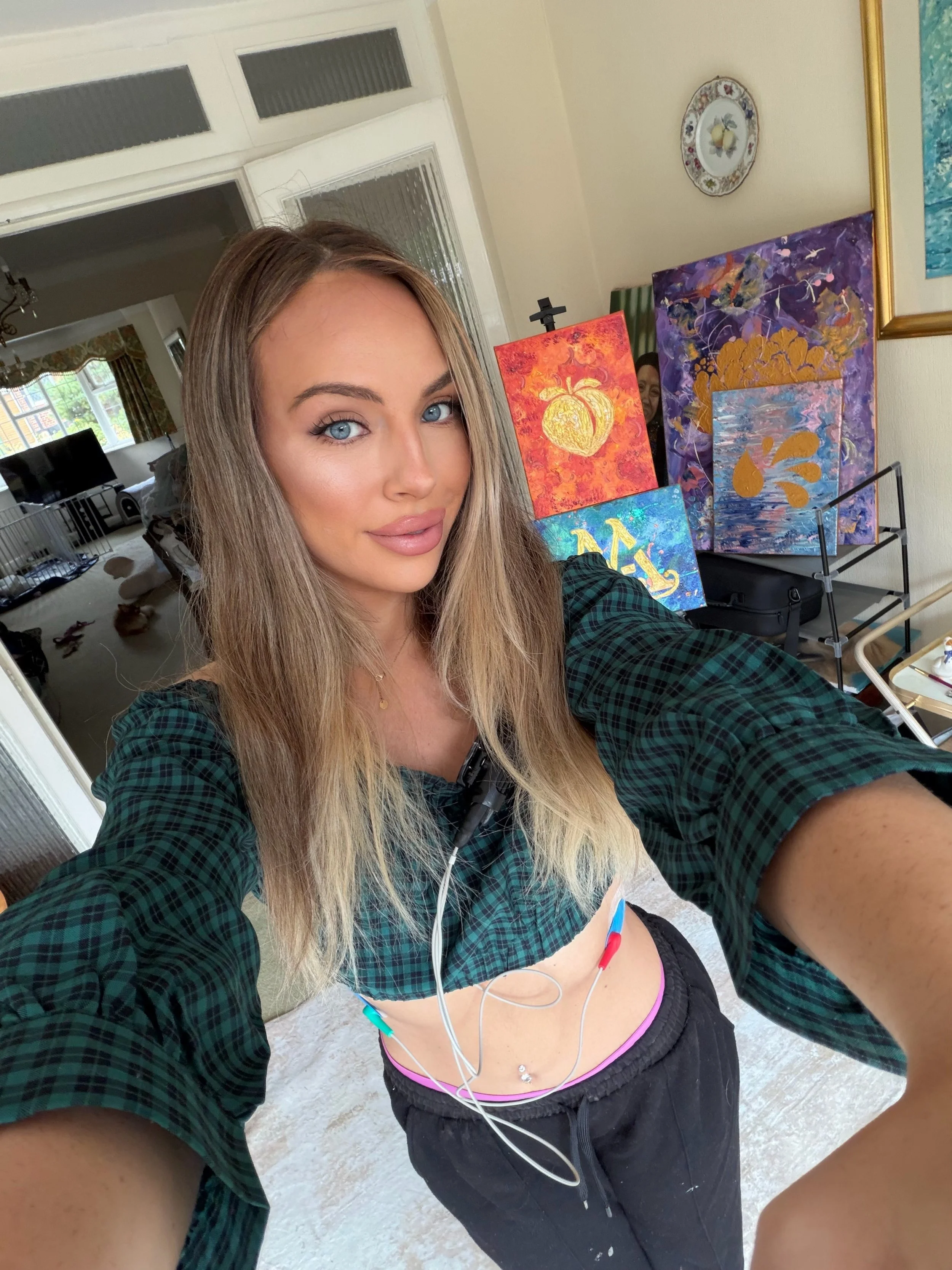 A woman with long blonde hair taking a selfie in a room with colorful abstract paintings on the wall, a window with curtains, and a glimpse of a living room with a dog on the floor.