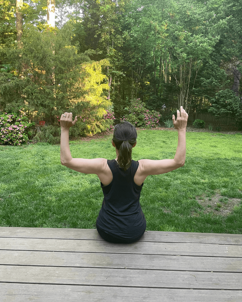 A woman sitting on a wooden deck with her back to the camera, flexing her arms in front of a lush green backyard with trees and flowering bushes.