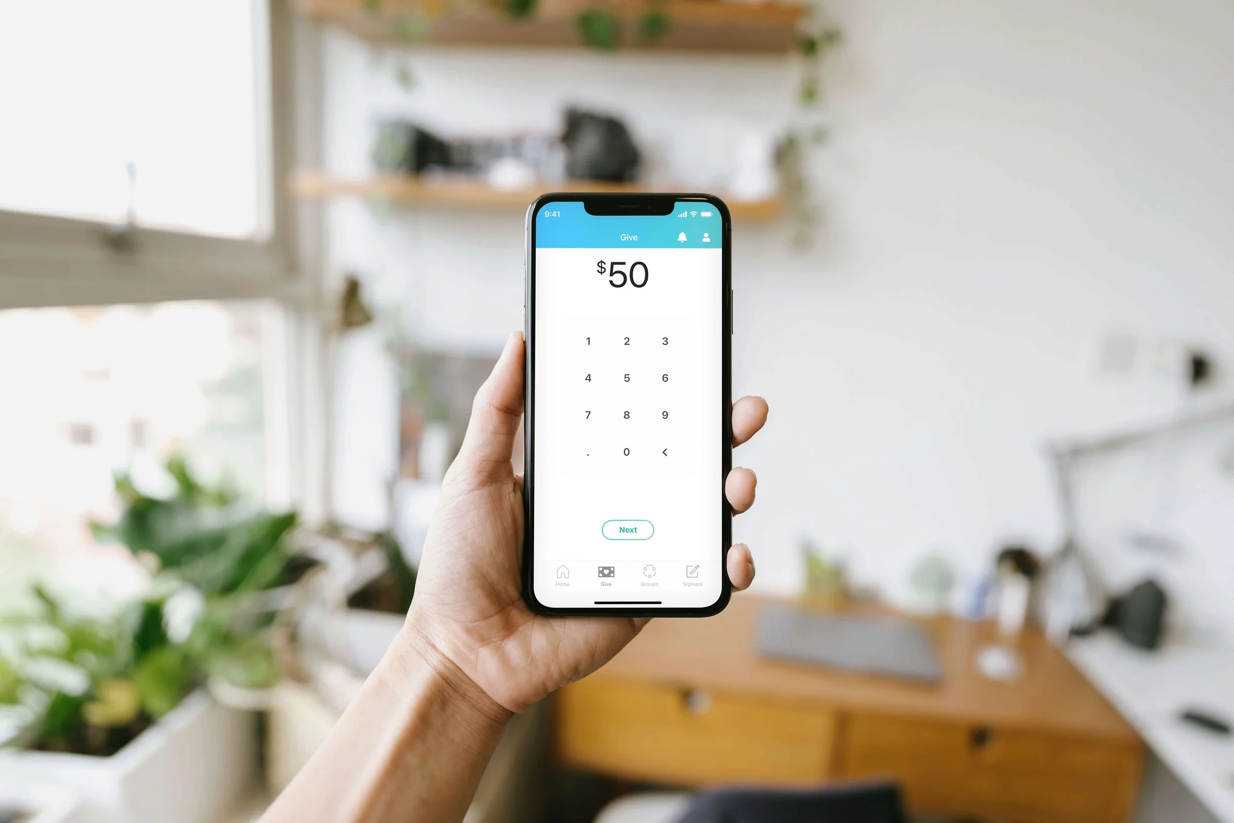 A person holding a smartphone displaying a donation screen with a $50 input on a digital keypad, in a well-lit room with plants and furniture in the background.