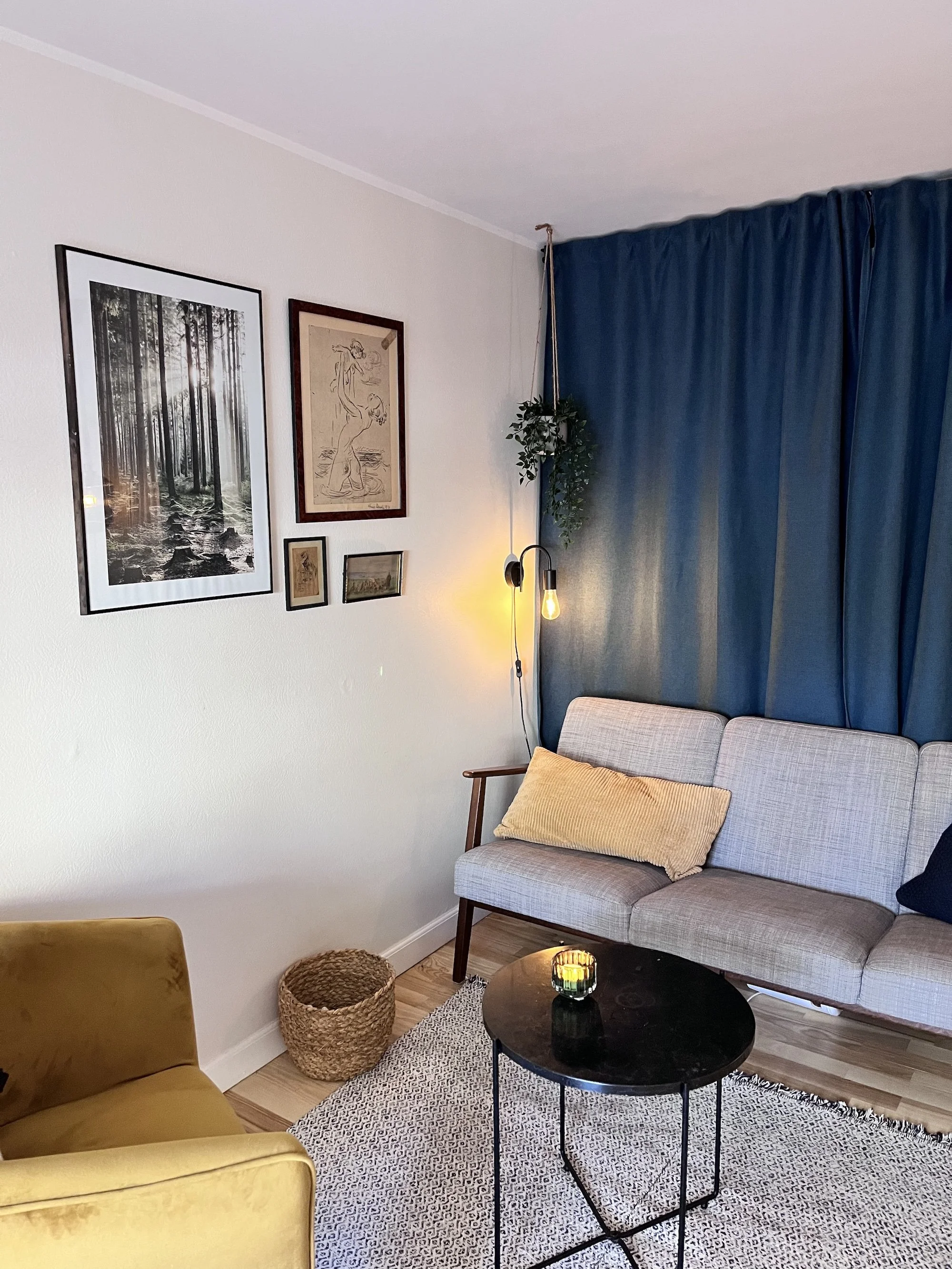 Stemningsfyldt terapilokale med blød sofa og stol, puder og varmt lys med kunst på væggen og grøn plante hængende fra loftet.
