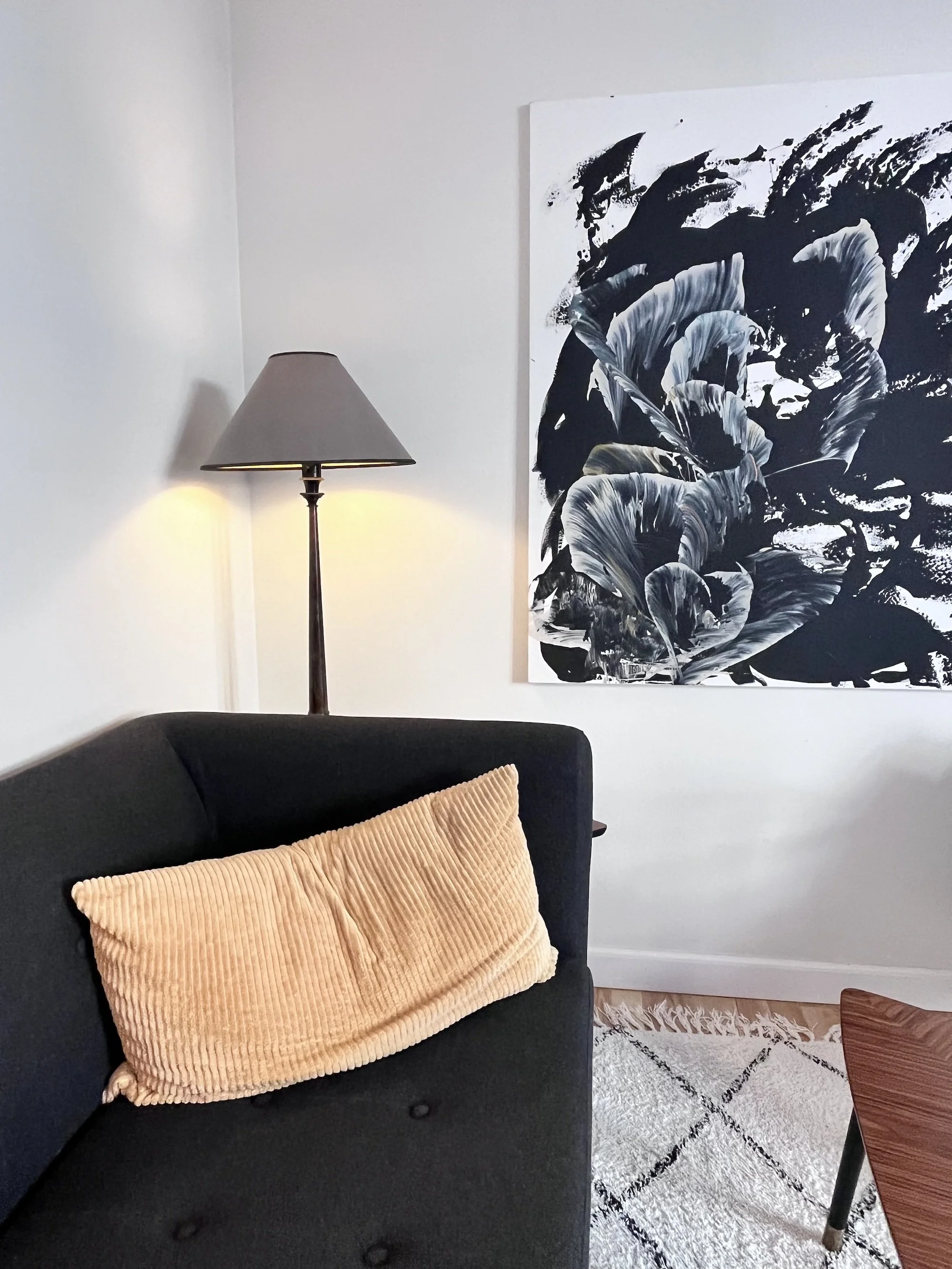 Venteområde i terapeutisk klinik. Mørk blød sofa med orange pude, varmt lys fra standerlampe, gulvtæppe, bord og et abstrakt maleri.