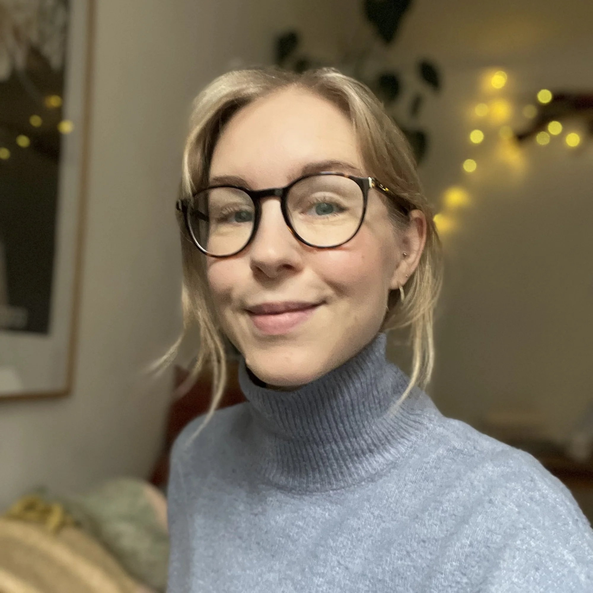 Portrait of therapist Siri Schwabe in a turtleneck sweater with blonde hair and glasses looking into the camera with a blanket, an art poster, a plant, and fairy lights in the background.