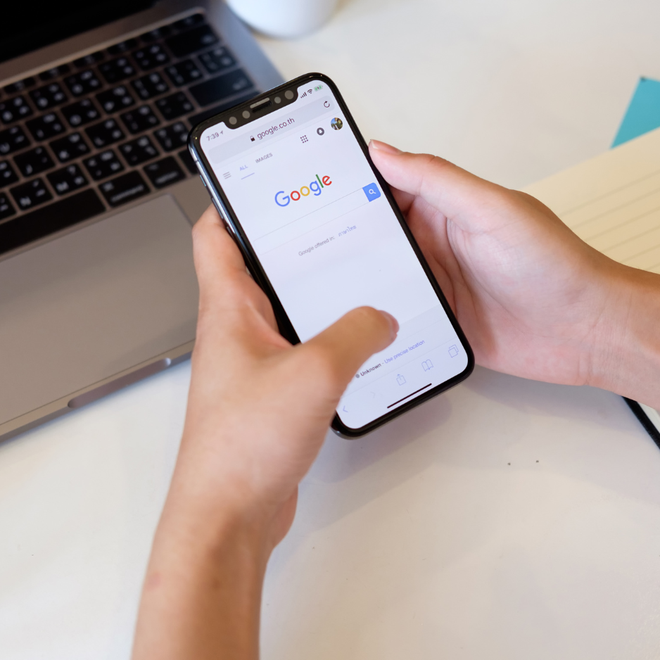Person holding a smartphone with Google search page on the screen, laptop on a white desk, partially visible notebook and blue sticky note.