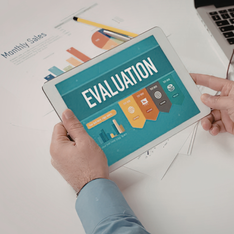 Person holding tablet displaying the word 'Evaluation' with business icons and bar graphs on a light blue background, on a desk with reports and a laptop.