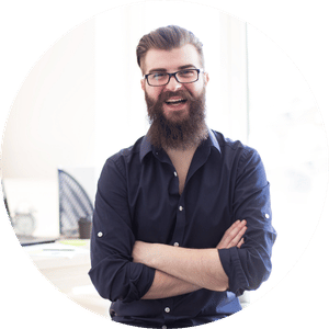 A smiling man with glasses and a beard, wearing a dark shirt, standing with arms crossed in an office environment.