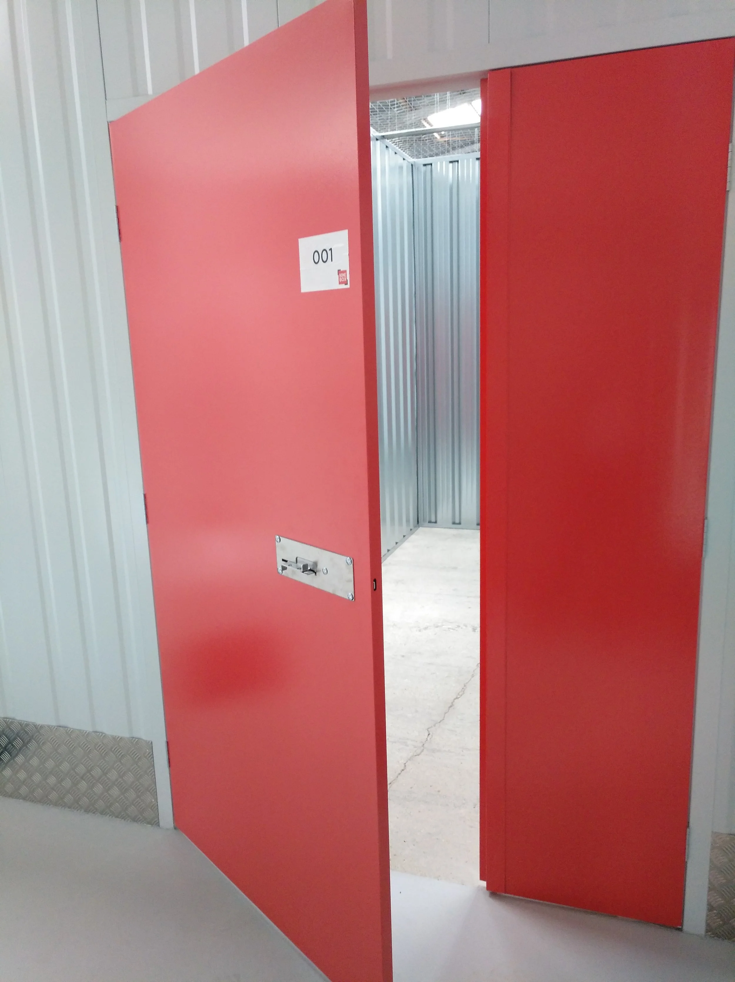 Puerta de un casillero de color rojo parcialmente abierta en un entorno industrial o de almacenamiento con paredes de paneles metálicos y suelo de concreto.